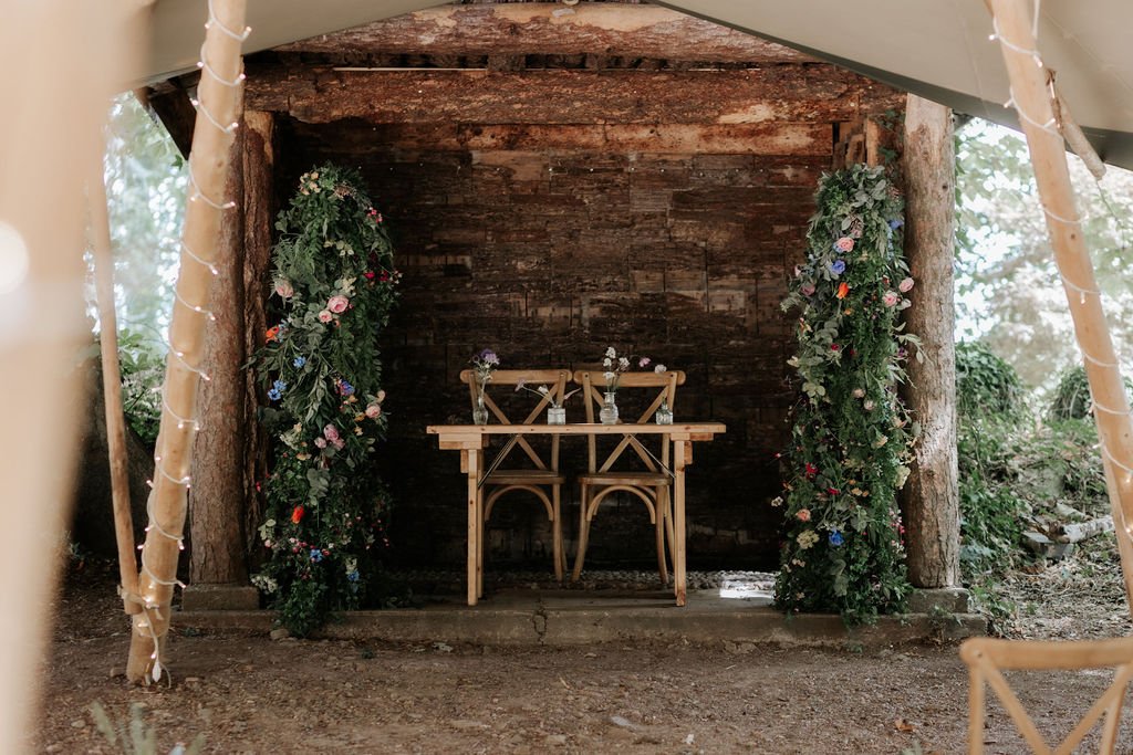 Two artificial floral pillars framing the rustic wooden ceremony shelter at Tipis at Whatton House.