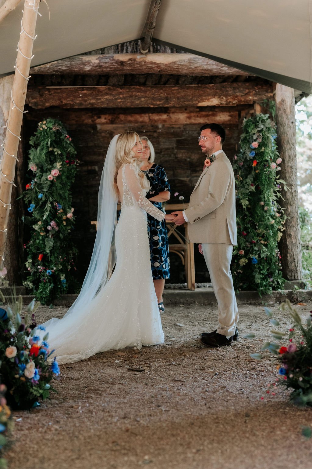 Couple exchanging vows between colourful artificial floral pillars at the wooden ceremony shelter at Tipis at Whatton House.