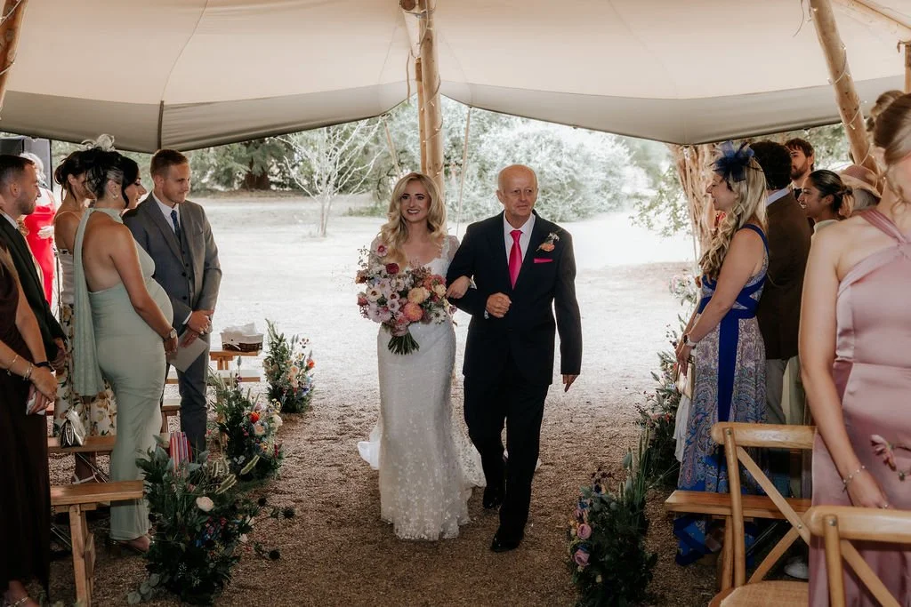 Bride walking down the outdoor ceremony aisle beside colourful artificial flower arrangements at Tipis at Whatton House.