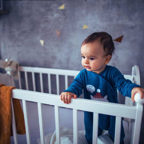 A young child standing in a white crib, holding onto the crib rails, wearing a blue pajamas with a cartoon character and the word 'Save' on it, in a nursery with a gray wall and decorative paper triangles.