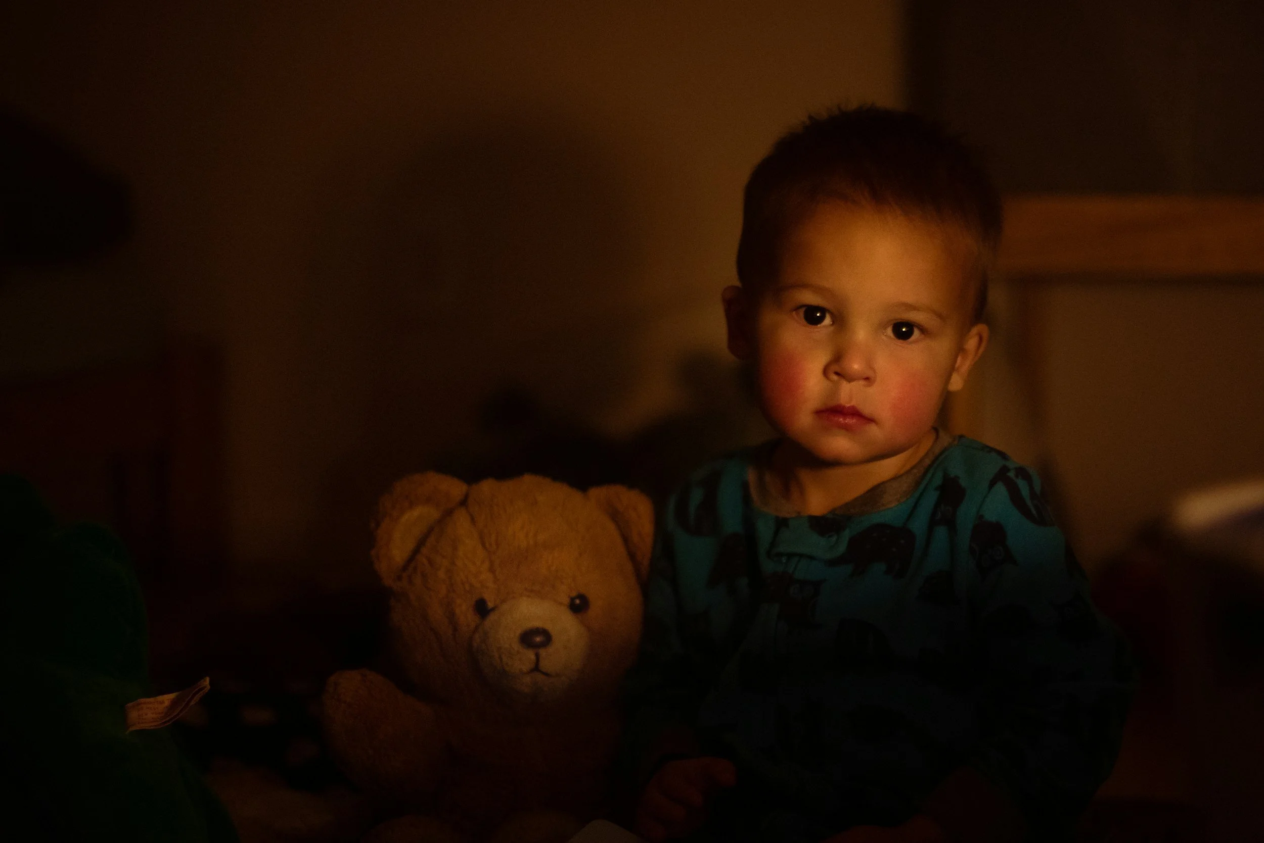 A young child with short hair and a serious expression, sitting next to a brown teddy bear, in a dimly lit room.