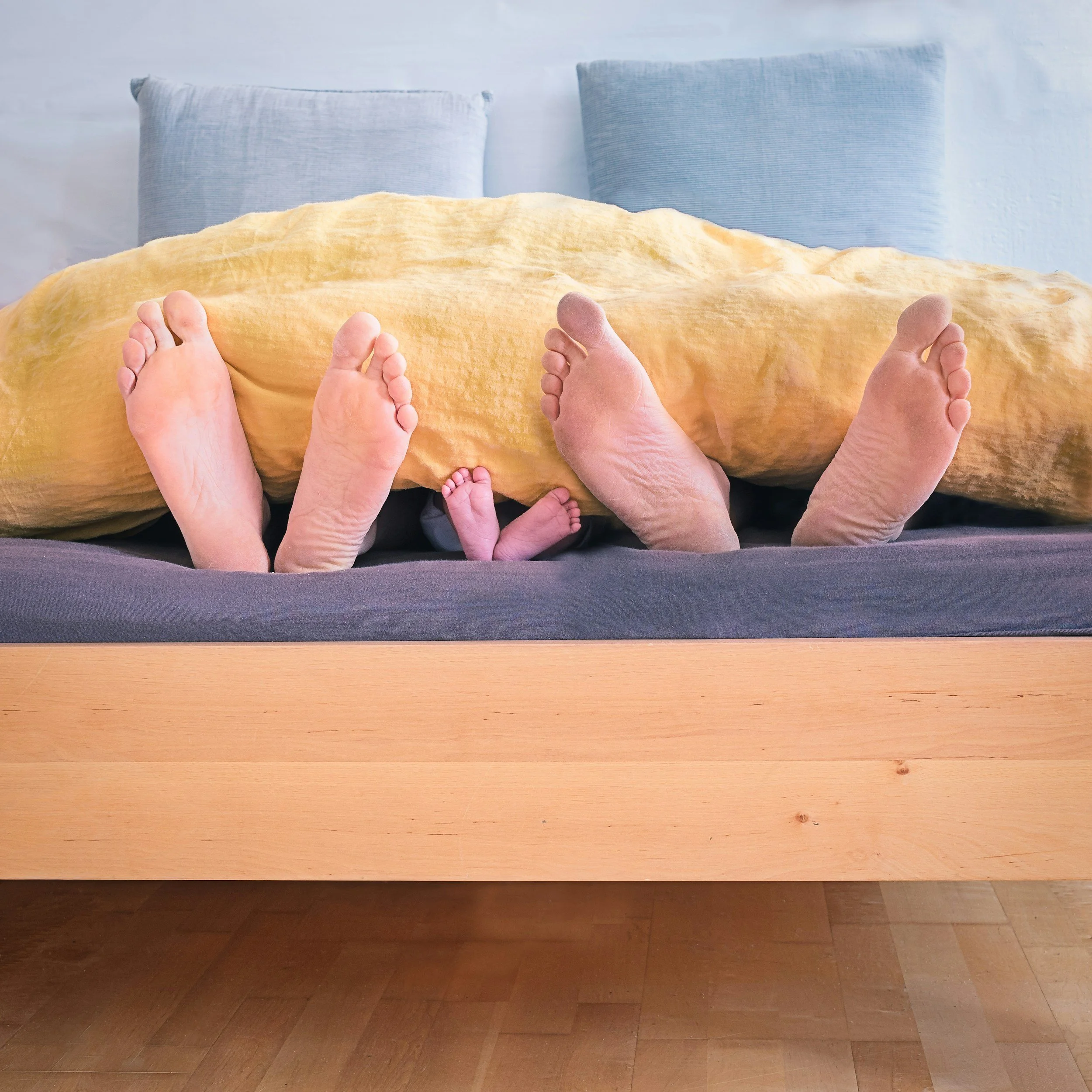 Multiple pairs of feet of different ages and sizes peeking out from under a yellow blanket on a bed with blue pillows.