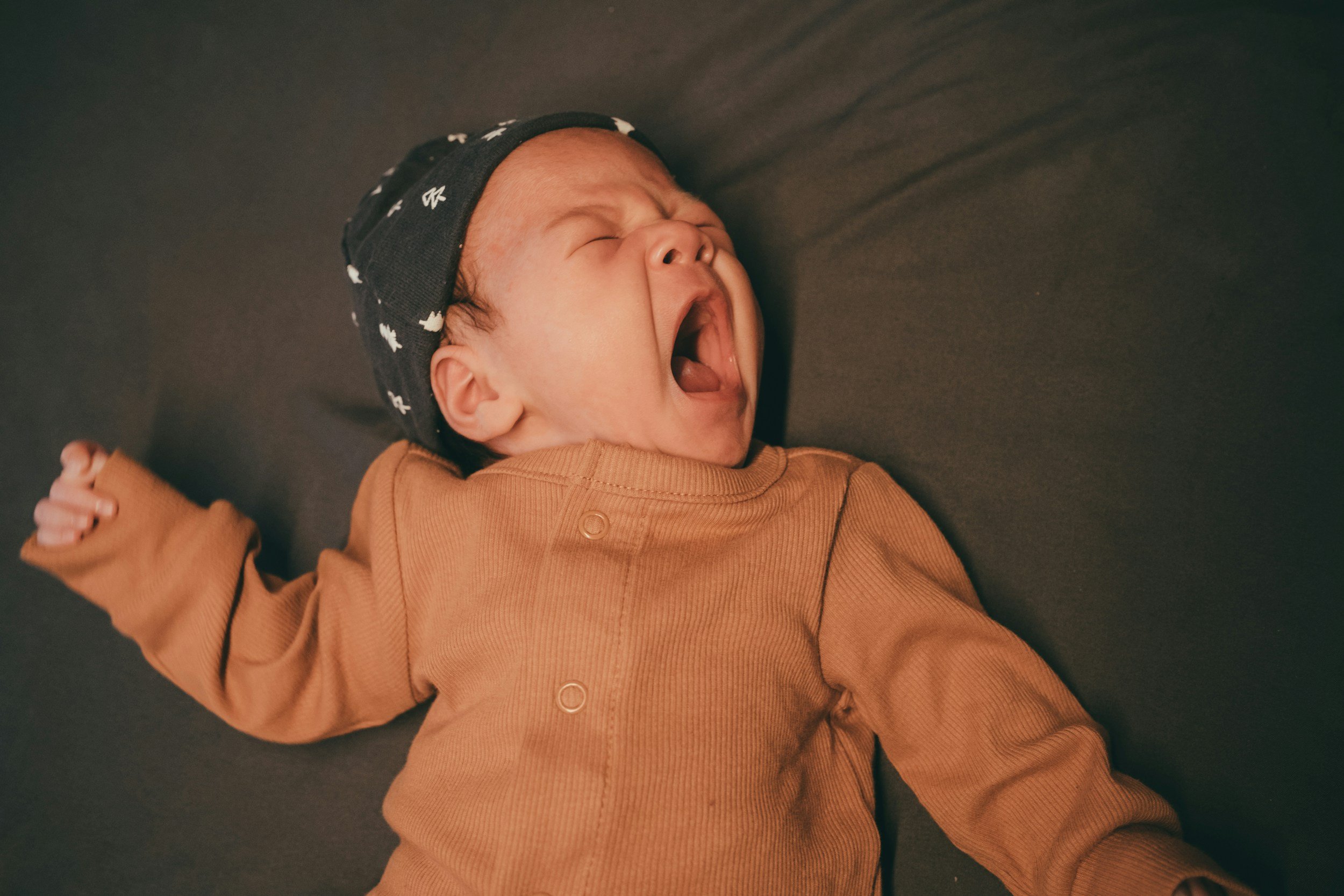 A baby lying on a dark surface, yawning with eyes closed and mouth wide open. The baby is wearing a black headband with white patterns and a brown long-sleeve shirt.