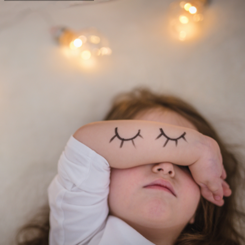 A young girl lying down with her eyes covered by her arm, which has a cartoon eyelash design drawn on it, suggesting she is sleeping peacefully. Soft, warm lights are in the background.