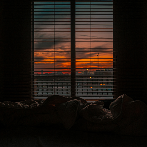 View through window blinds of a colorful sunset over a city skyline at dusk.