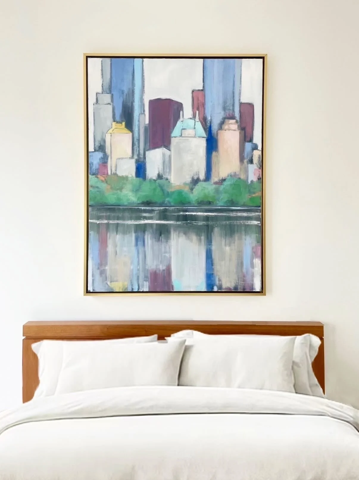 Interior of a bedroom with a wooden headboard, white bedding, and a framed painting of a city skyline with skyscrapers reflected in water above the bed.