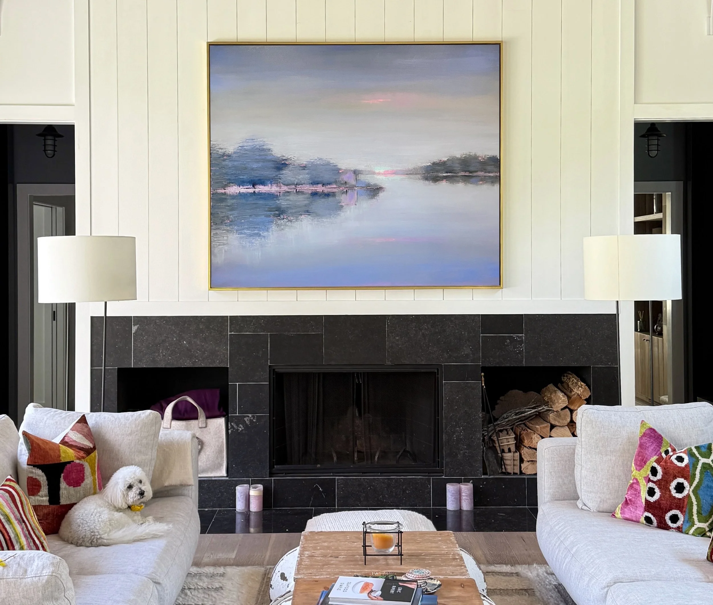Living room with a large landscape painting above the fireplace, beige sofas with colorful pillows, a small white dog on one sofa, coffee table with books and candles, log storage beside the fireplace.