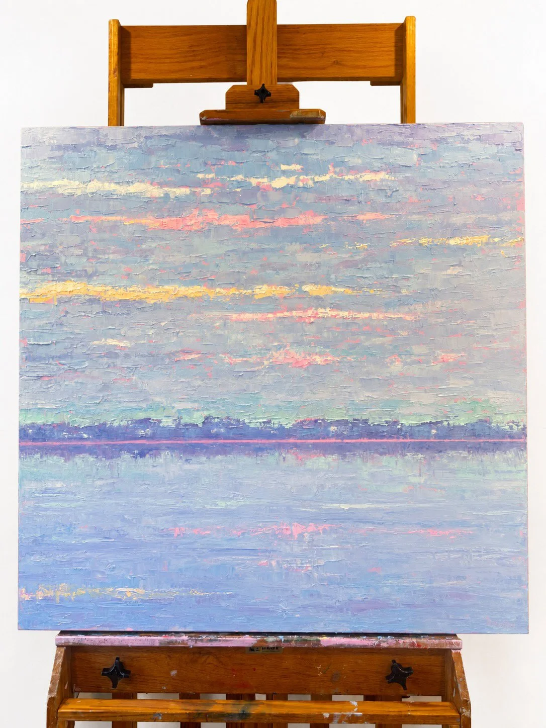 A pastel-colored landscape painting on an easel, depicting a calm body of water and a colorful sky with clouds, silhouetted land in the distance.