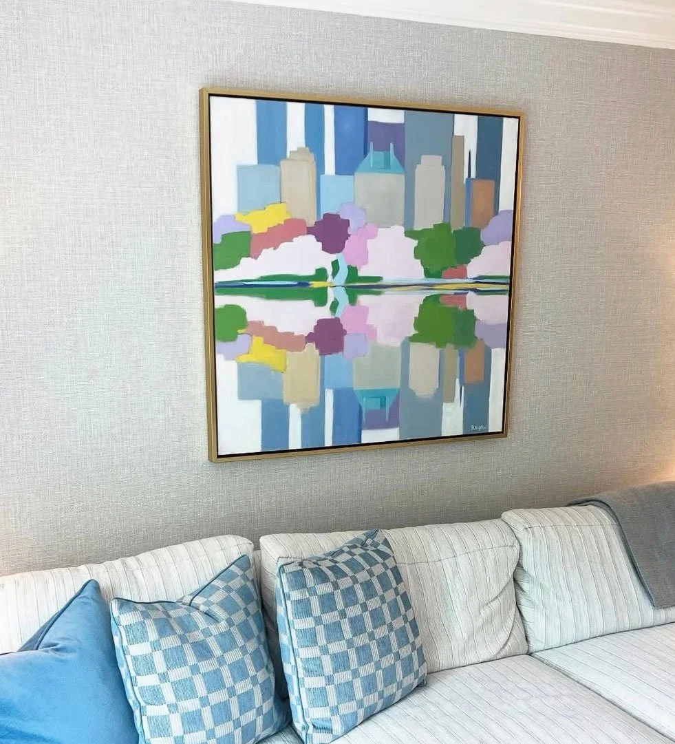 Colorful abstract painting of a city skyline with buildings and trees reflected in water above a white couch with blue and checkered pillows.
