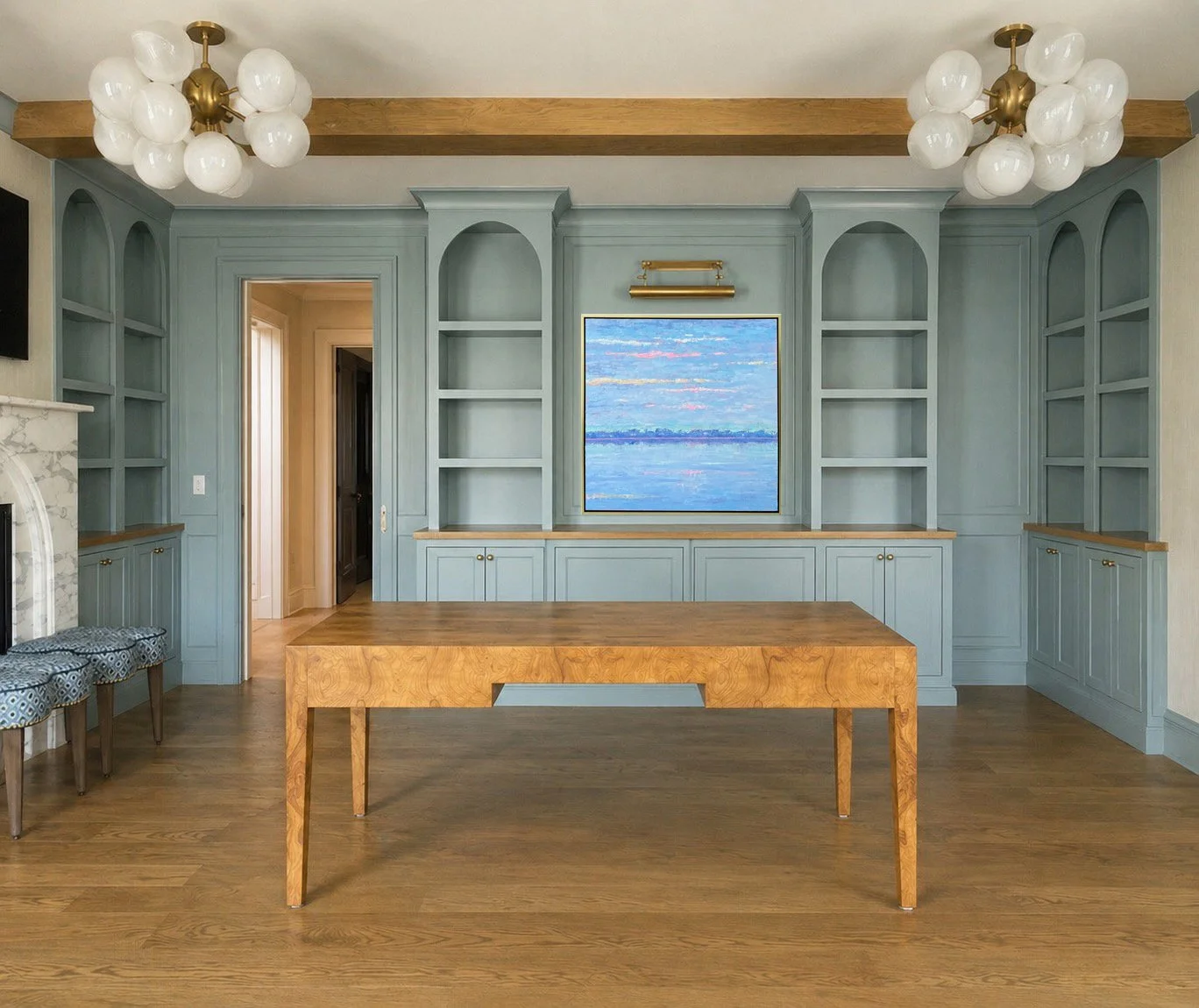 Living room with built-in blue bookshelves, a large window with a landscape painting, a wooden coffee table, hardwood floors, and two chandeliers with globular white lights.
