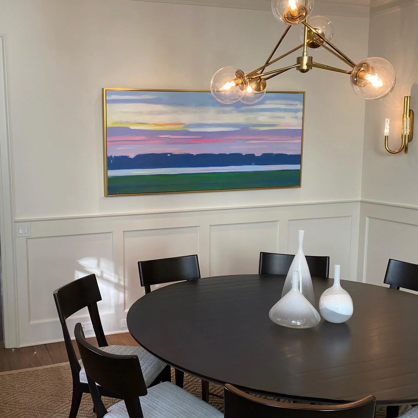 A dining room with white paneled walls, a large landscape painting, a modern chandelier with multiple glass globes, a dark wooden round table, and black chairs. Three white vases are on the table.