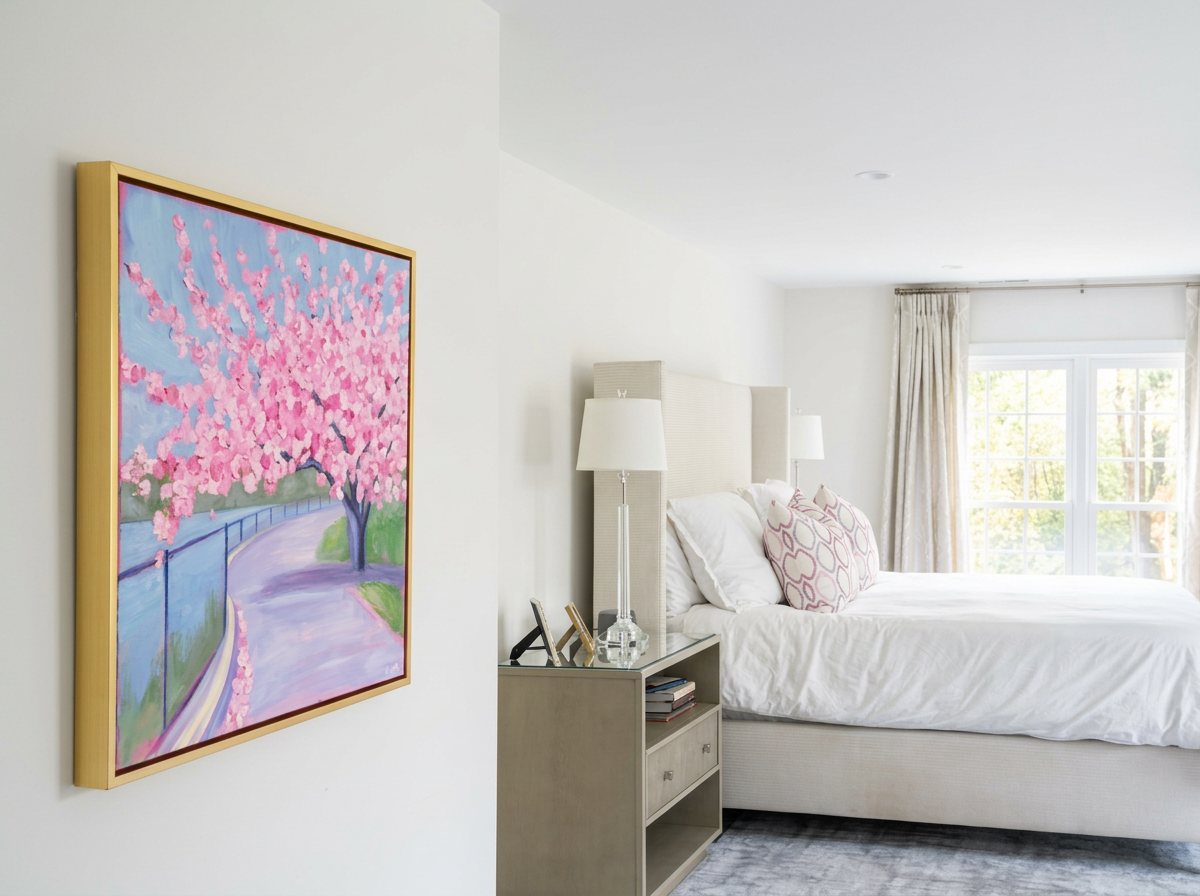 A bedroom with a white bed, pink and white pillows, a nightstand with picture frames, lamps, and books, and a large window with curtains showing trees outside.