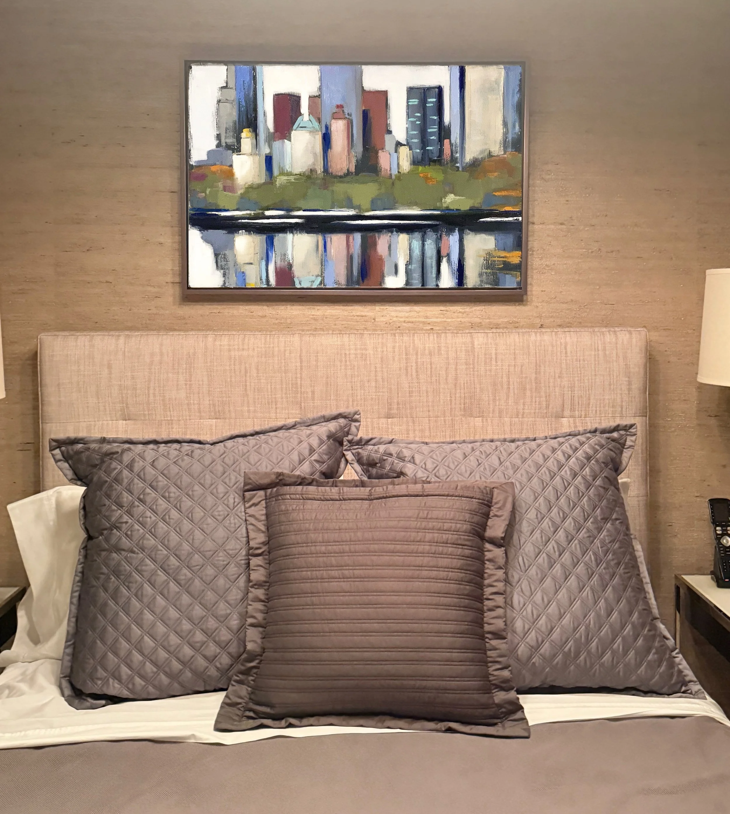 Hotel bedroom with a bed, beige headboard, taupe pillows, white sheets, and a framed cityscape painting of skyscrapers above the bed.