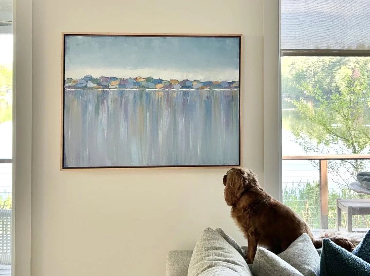 A living room with a large abstract landscape painting on the wall and a brown dog sitting on a gray sofa, looking out the window.