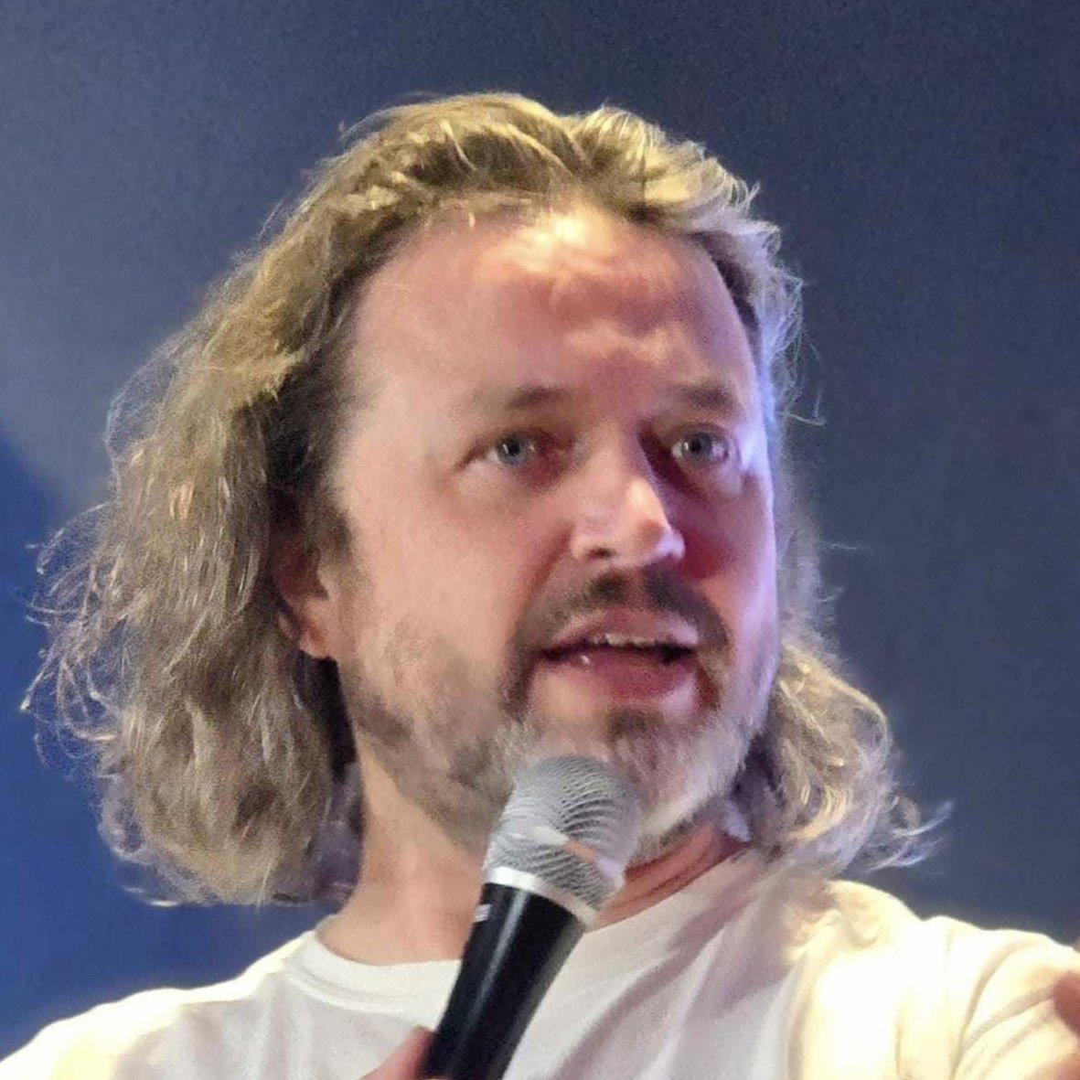 A man with long, wavy, blond hair, and a beard, holding a microphone and speaking.