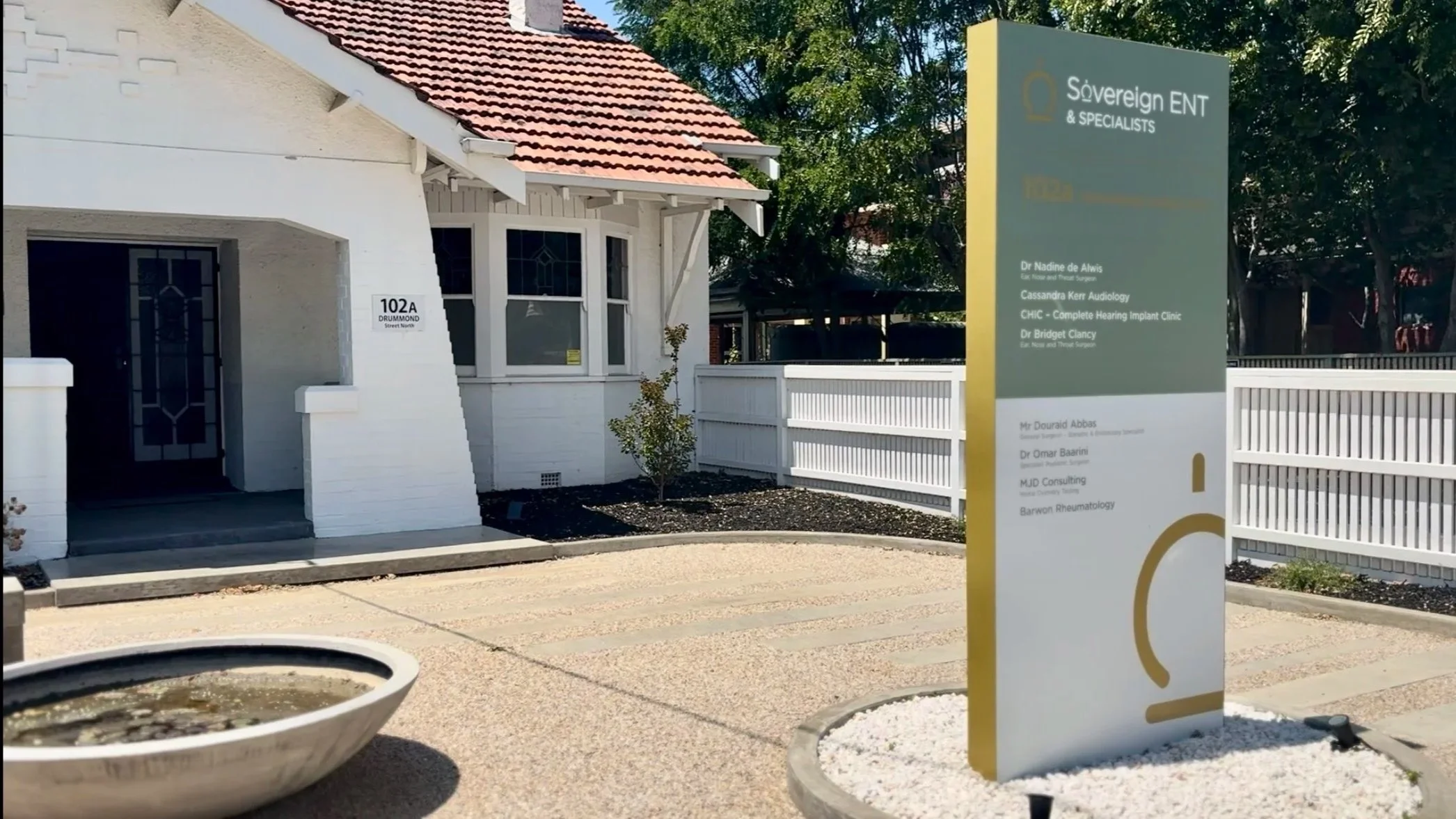 Exterior of a white building with a red tiled roof and a sign that reads 102A Drummond Street North. There is a large sign outside for Sovereign ENT & Specialists, listing names and services, surrounded by a white fence and landscaping with a small tree and white decorative rocks.