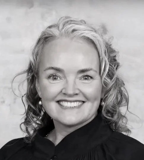 Black and white photo of a smiling woman with short hair, wearing a textured blazer and a necklace, sitting in front of a window with flowers outside.