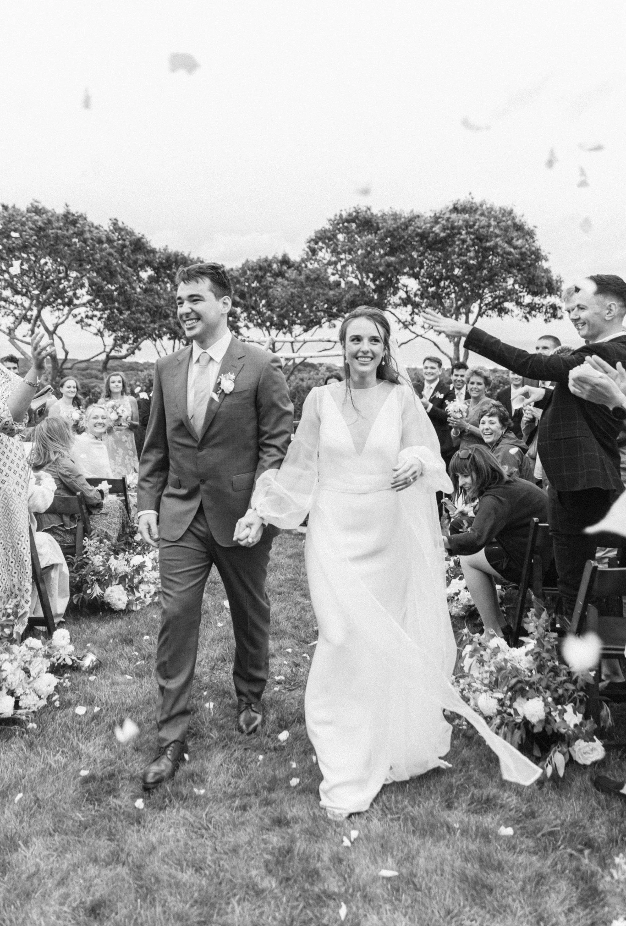 A cheerful bride and groom walk down the aisle after a beautiful outdoor ceremony at the grooms family home overlooking the Atlantic Ocean in Chilmark, on Martha's Vineyard. Flower petals fly through the air as guests cheer on with joy.