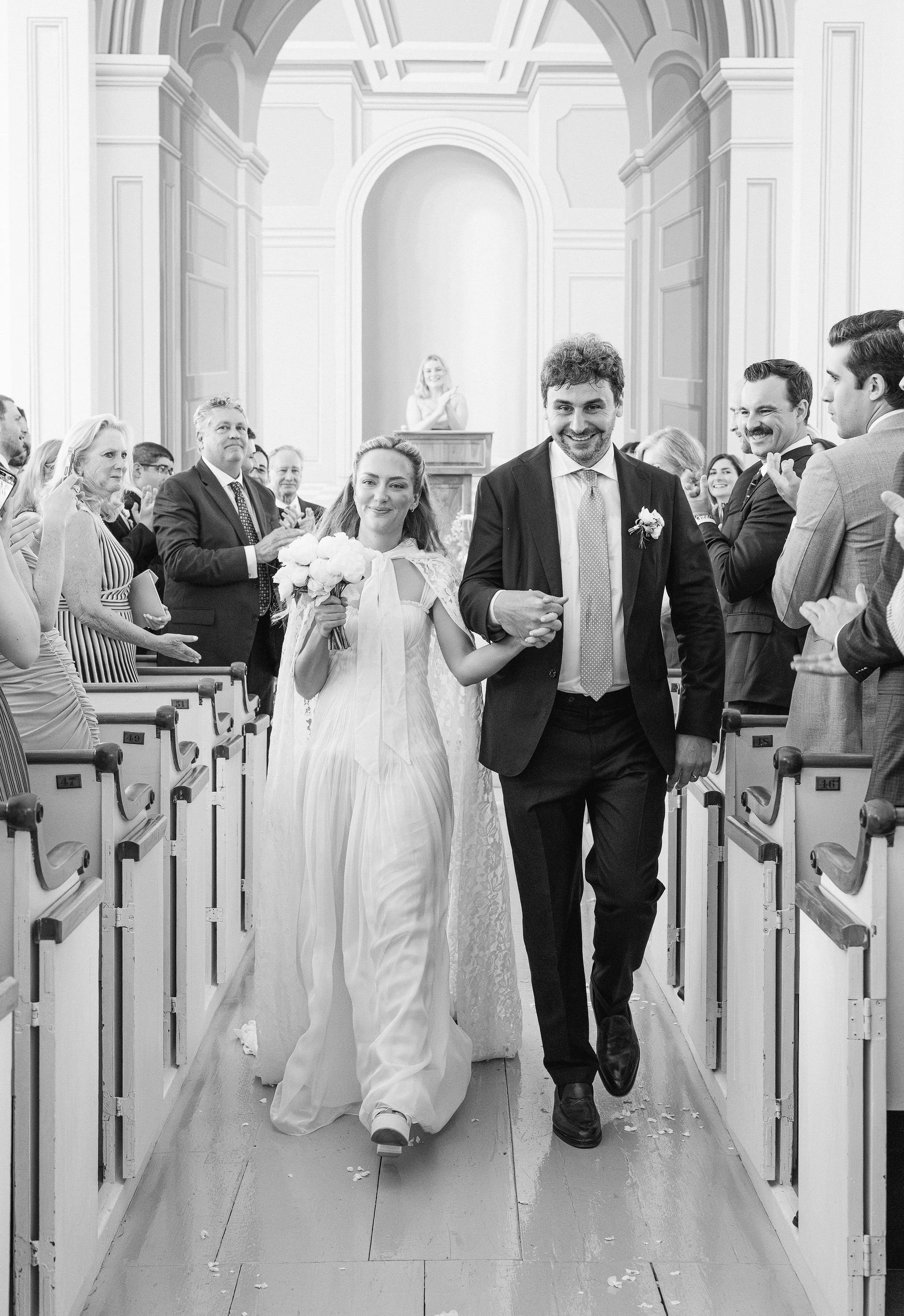 The beautiful backdrop of the Old Whaling Church in Edgartown on Martha's Vineyard frames this happy couple as they walk down the aisle after their ceremony. A tasteful, simple white bouquet and a chic couture dress and cape enhance the bride