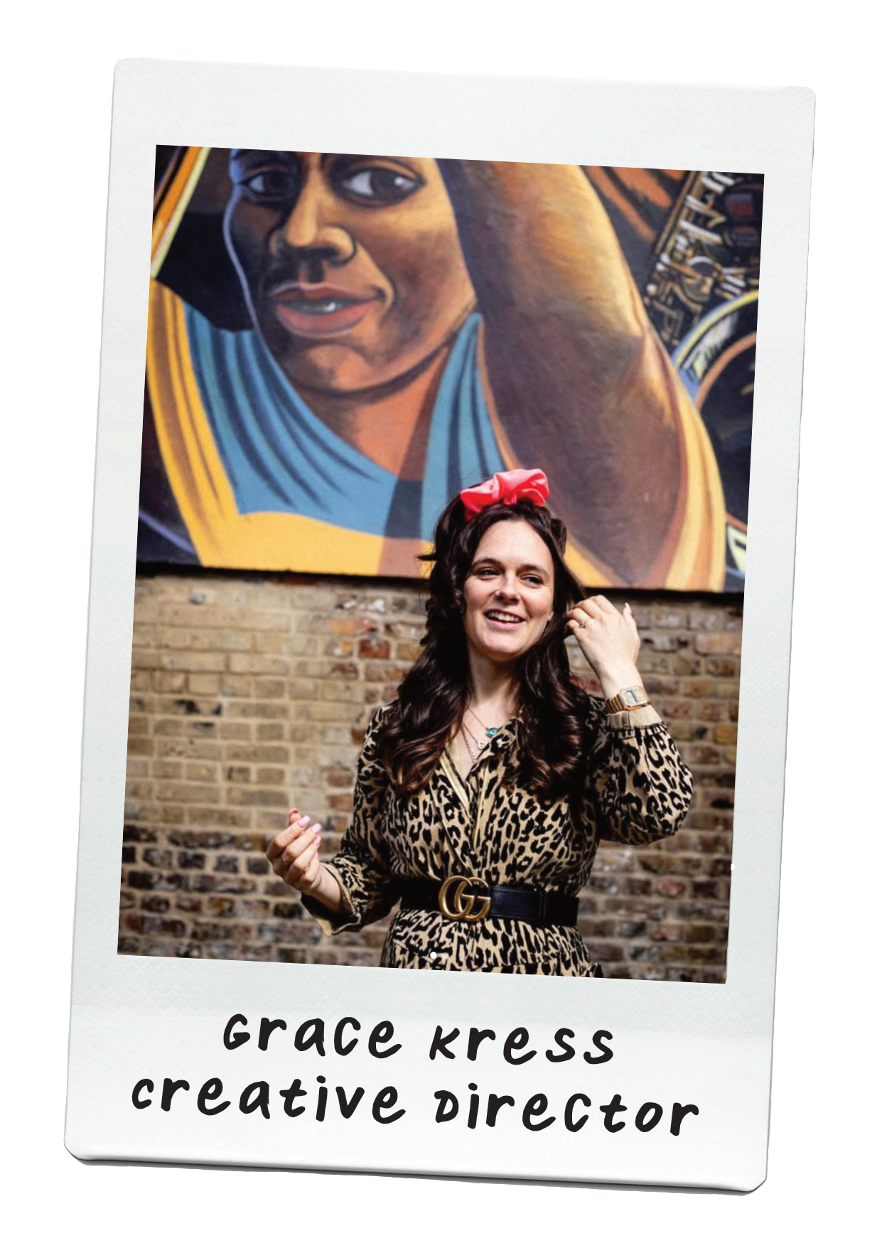 A woman with long brown hair, wearing a leopard print dress with a G belt and a pink bow headband, smiling and standing in front of a brick wall with a large mural of a woman in a blue shirt.