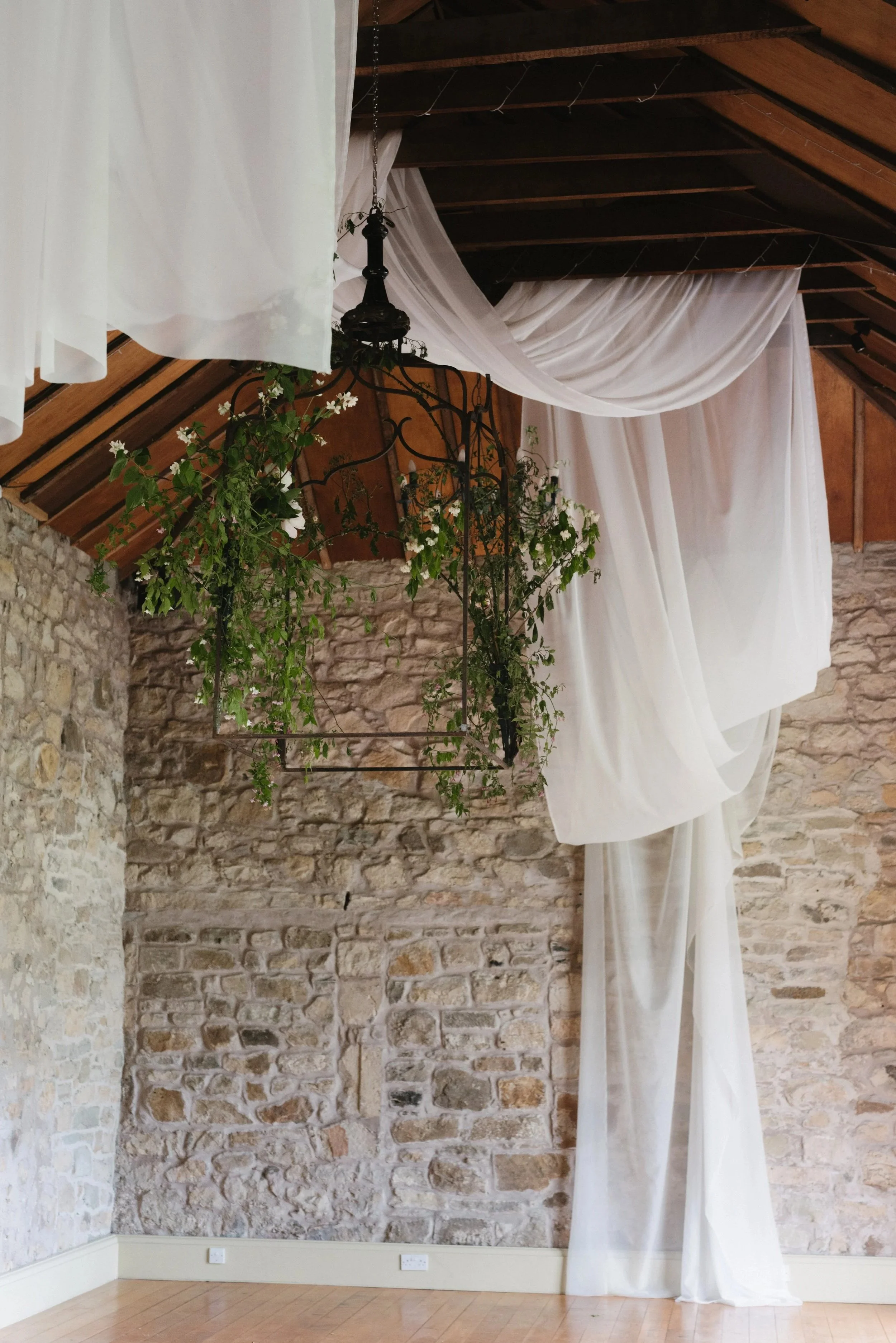 Interior of a rustic room with exposed stone wall, wooden ceiling, and white sheer curtains draped along the wall, with hanging greenery and a vintage chandelier.