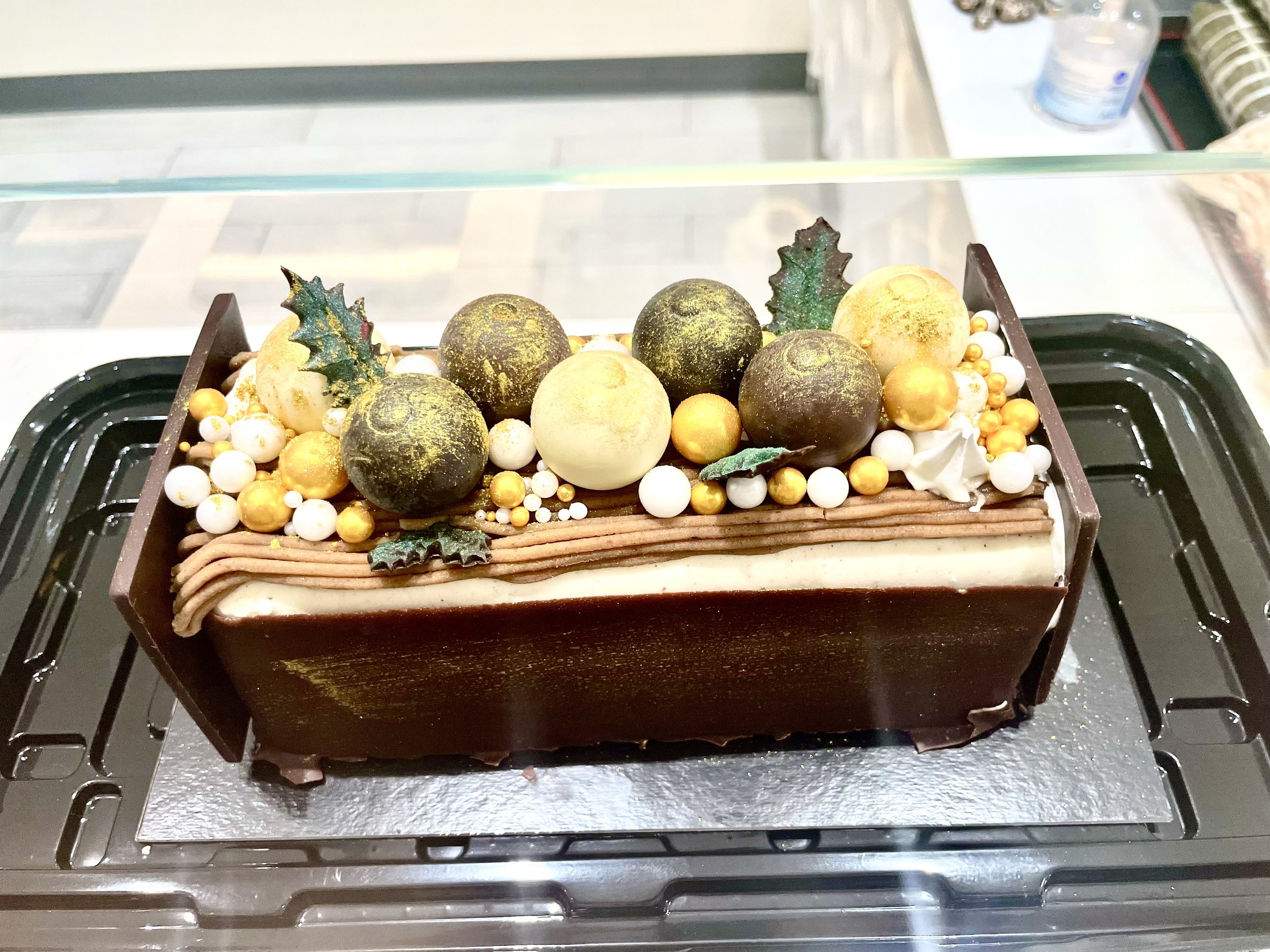 Decorative Christmas cake with gold and white spherical ornaments, holly leaves, and chocolate zigzag border.