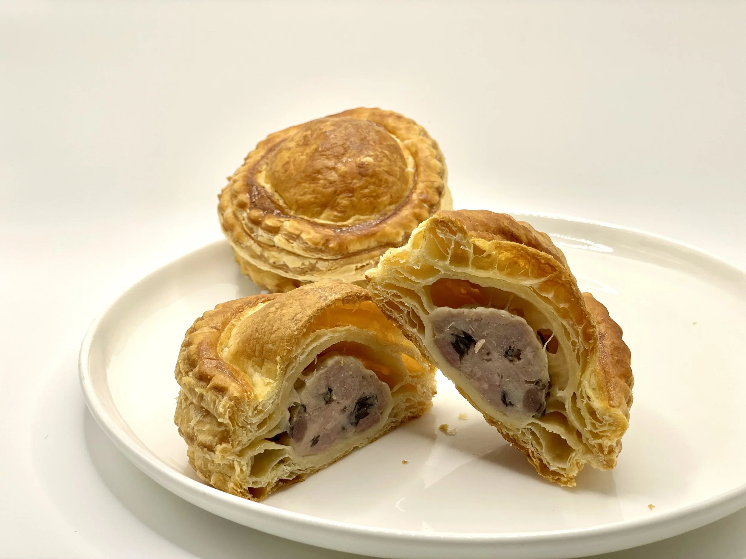 A white plate with sliced baked pastry containing sausage, cheese, and olives, with a whole pastry in the background.