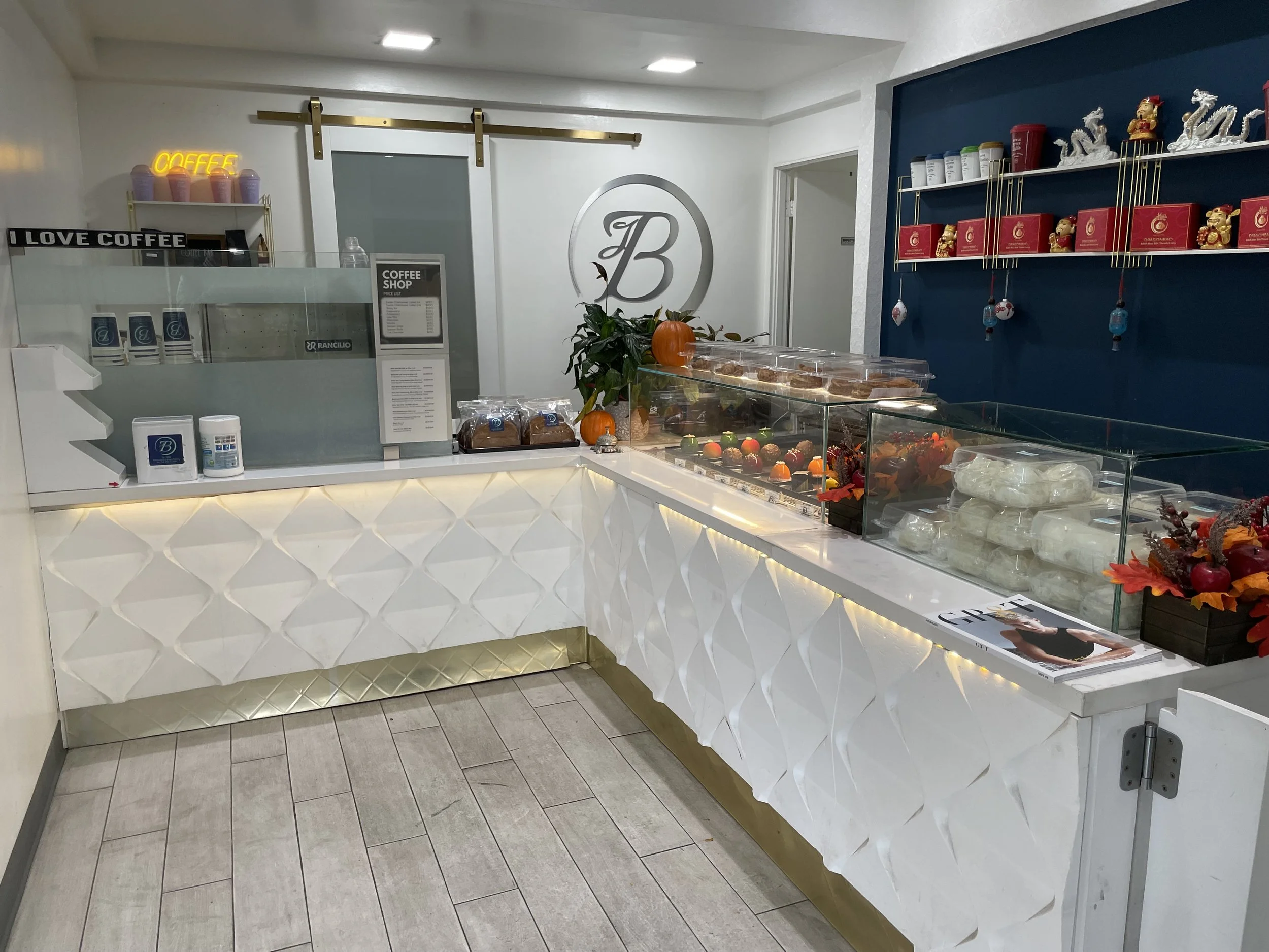 A cafe counter with a white front panel featuring quilted gold accents, displaying baked goods and desserts including cupcakes and pastries. Behind the counter, there is a small coffee shop sign, a menu, and a potted plant with pumpkins on top. To th