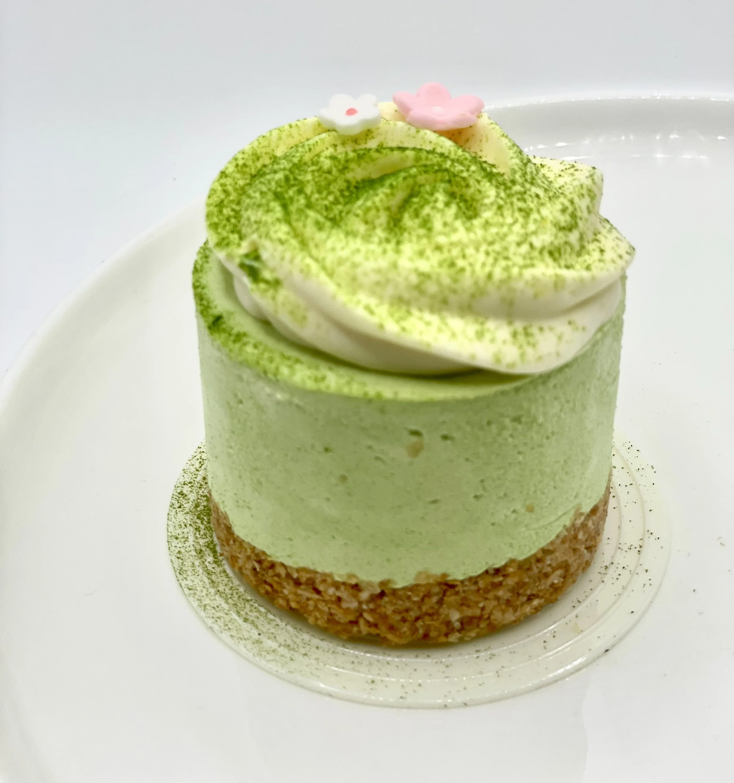 A small, round cake with white and green frosting, topped with pink and white fondant flowers, on a white plate with green sprinkles.