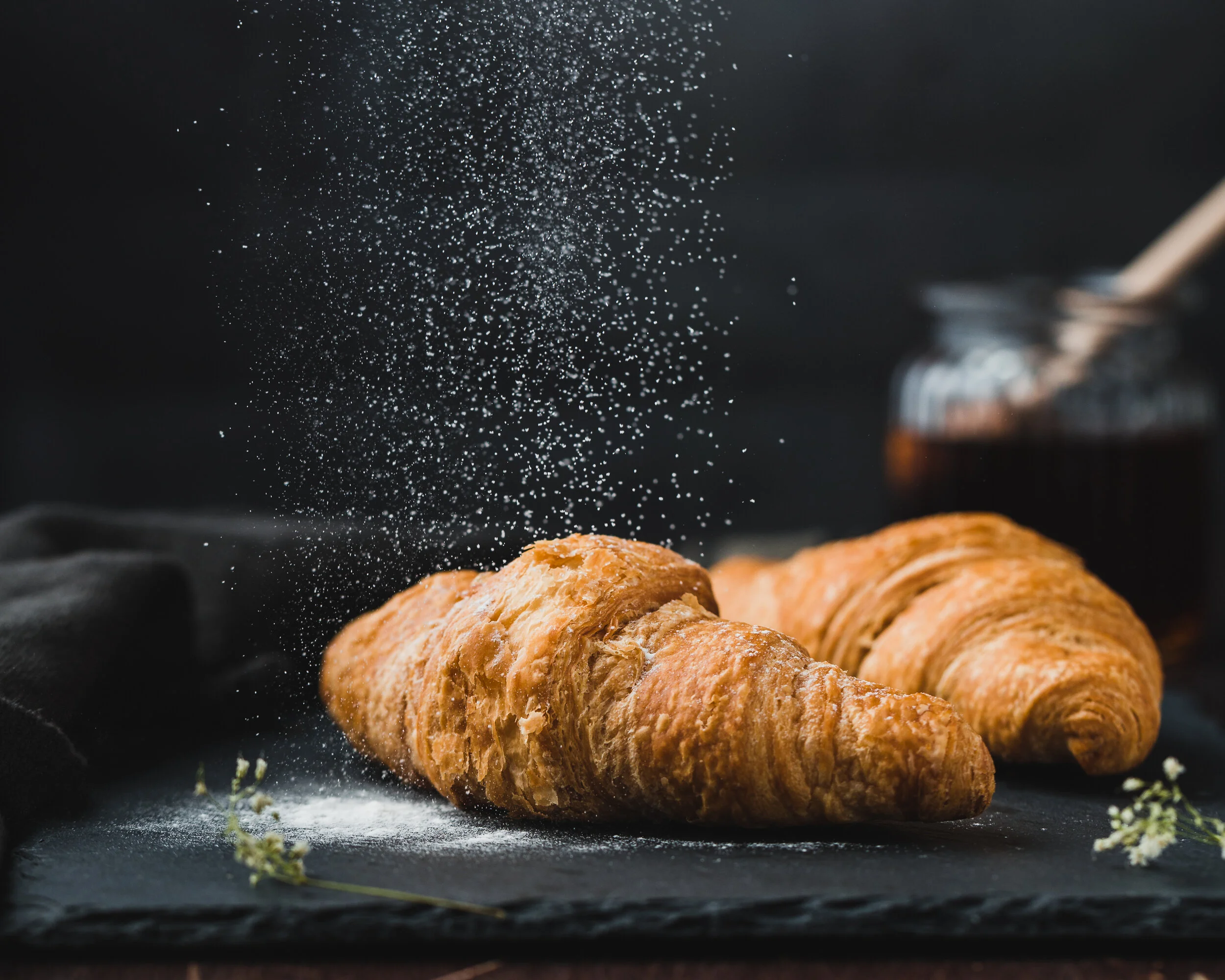 Two croissants on black parchment with sugar being sprinkled over them, jar of jam and a spoon in the background.