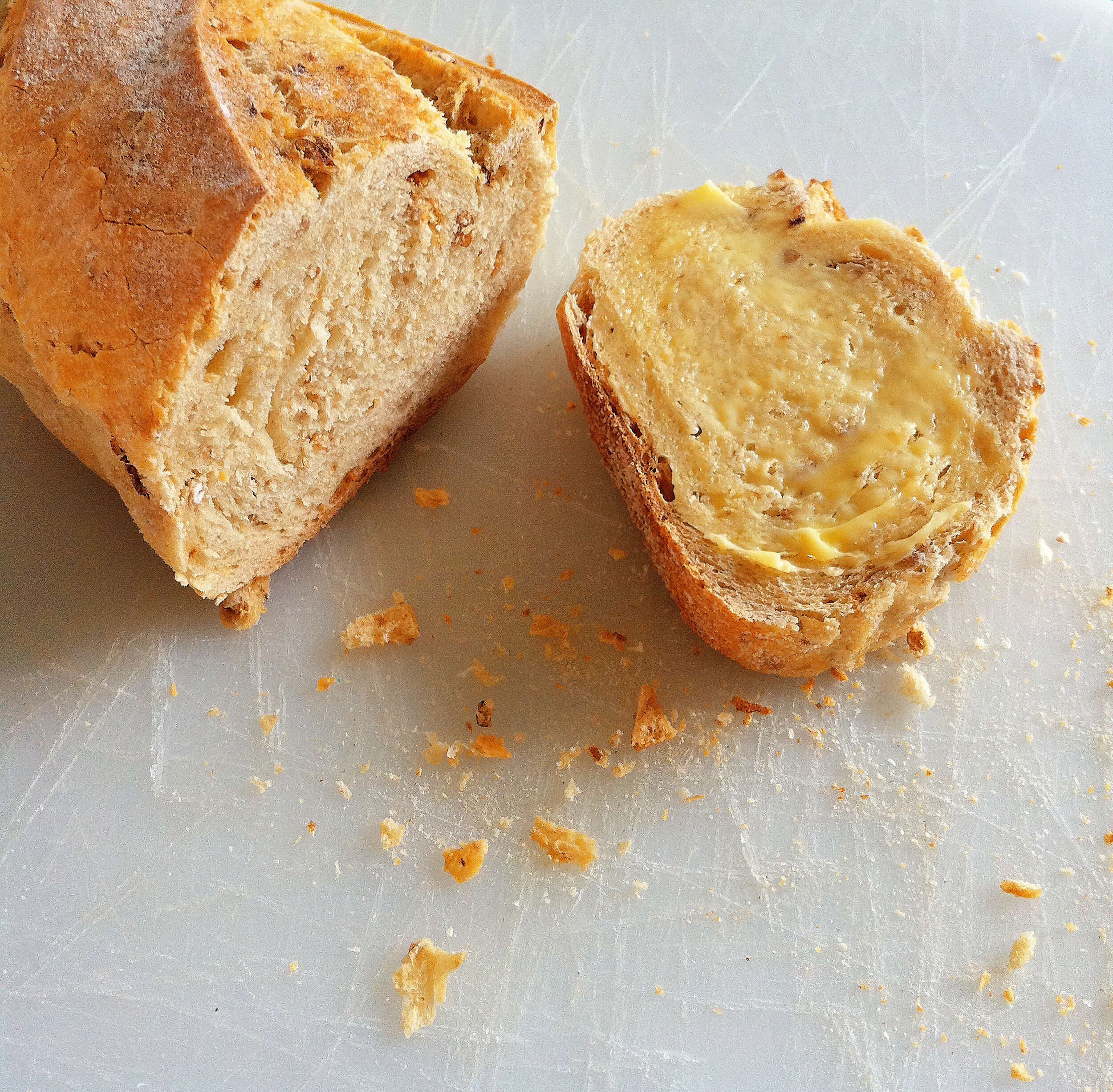 A loaf of bread with a slice cut off, buttered on top, on a white cutting board with crumbs around.
