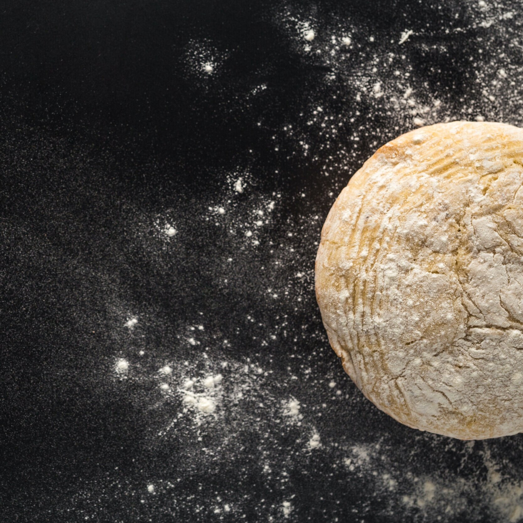 A ball of dough on a black surface, dusted with flour, with scattered flour particles around.