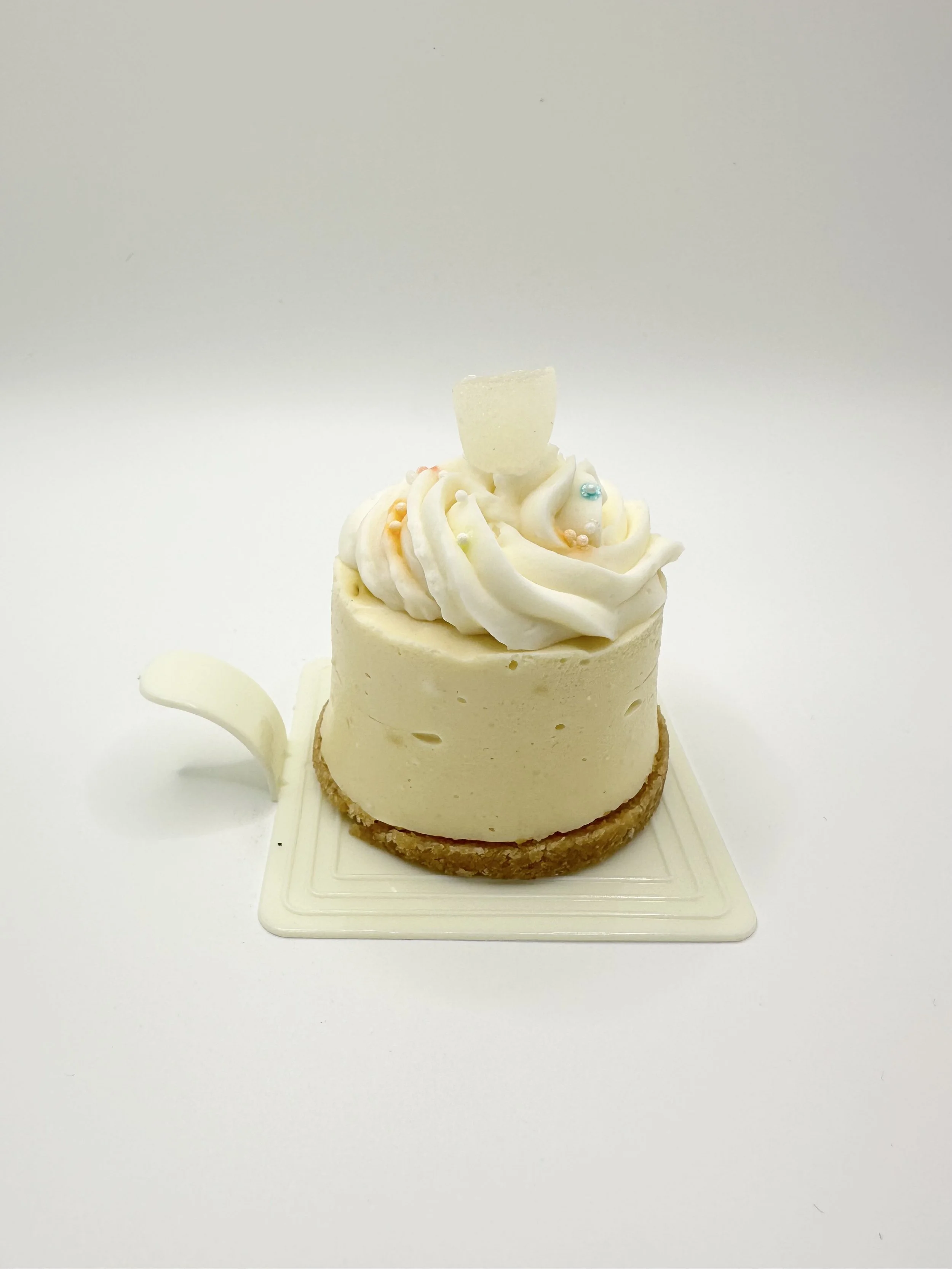 A dessert with a light-colored, round base, topped with white frosting piped in swirls, decorated with small colorful pearls, a white chocolate piece, and a white chocolate teapot on a white surface against a white background.