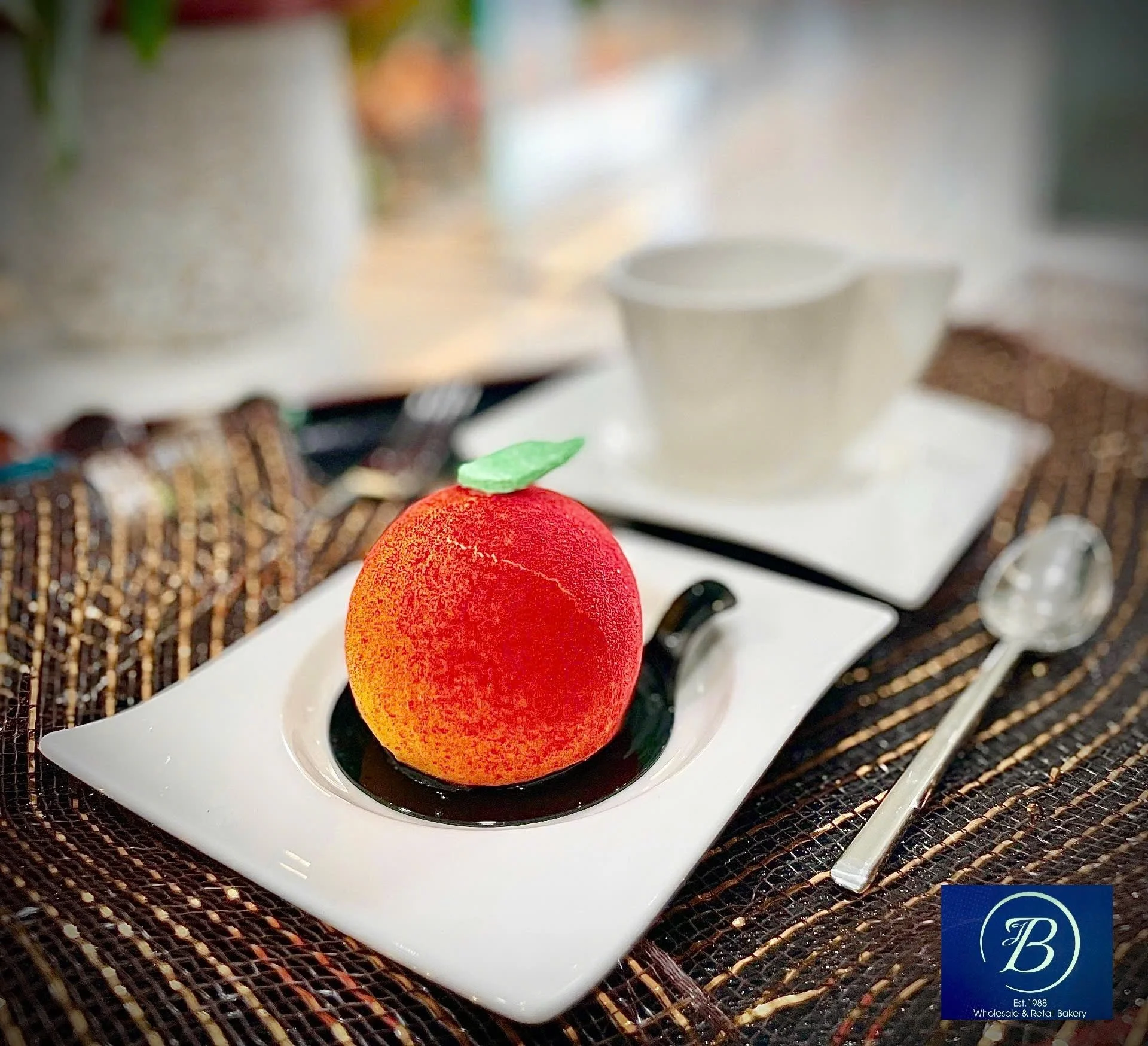 A spherical red dessert with a textured surface, topped with a small green leaf, sitting on a white rectangular plate with sauce, on a woven placemat.