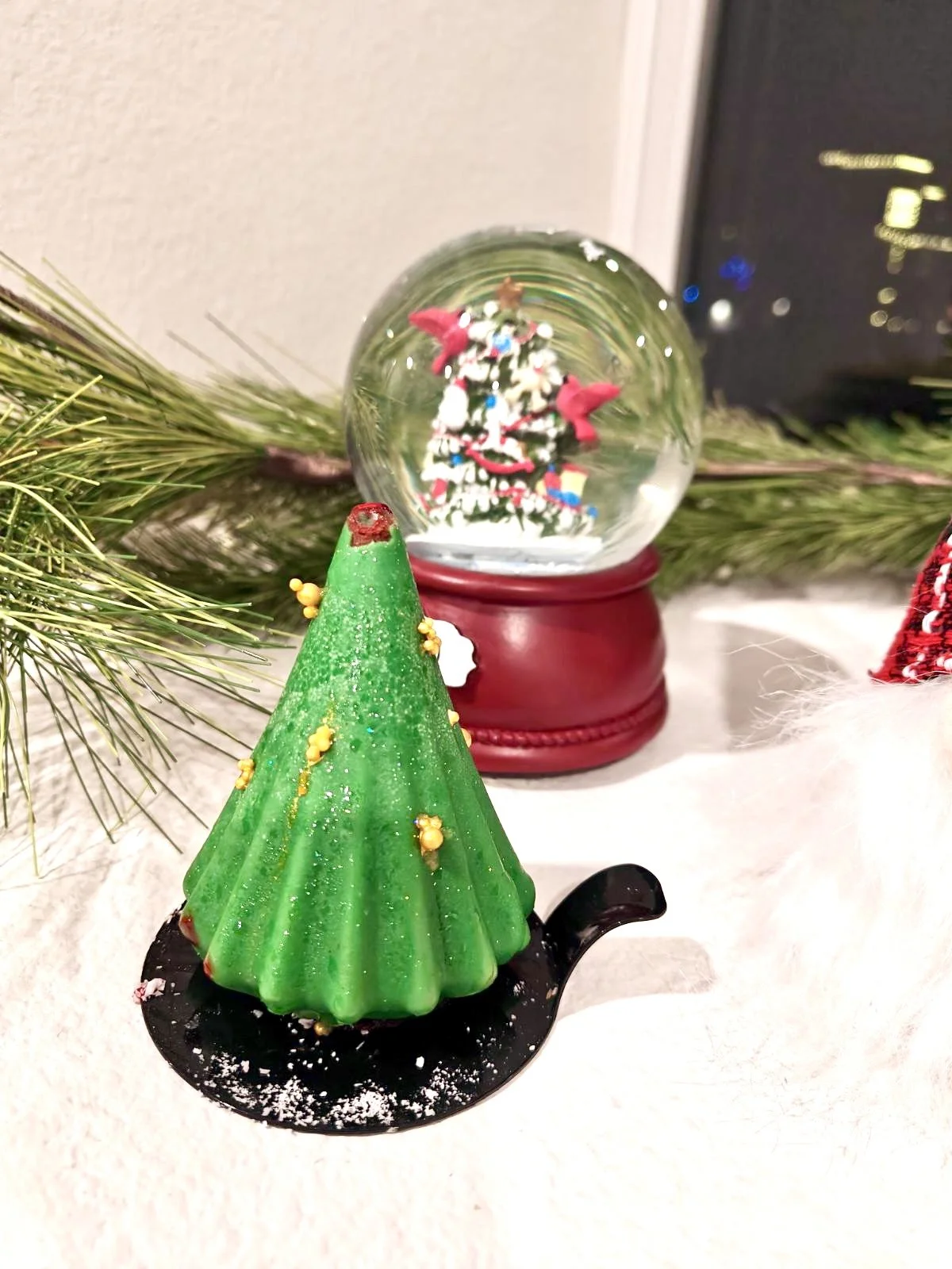 A Christmas-themed scene featuring a small green tree-shaped object with yellow decorations on a black holder, a snow globe with a Christmas tree inside on a red base, and a sprig of pine, all on a white surface.