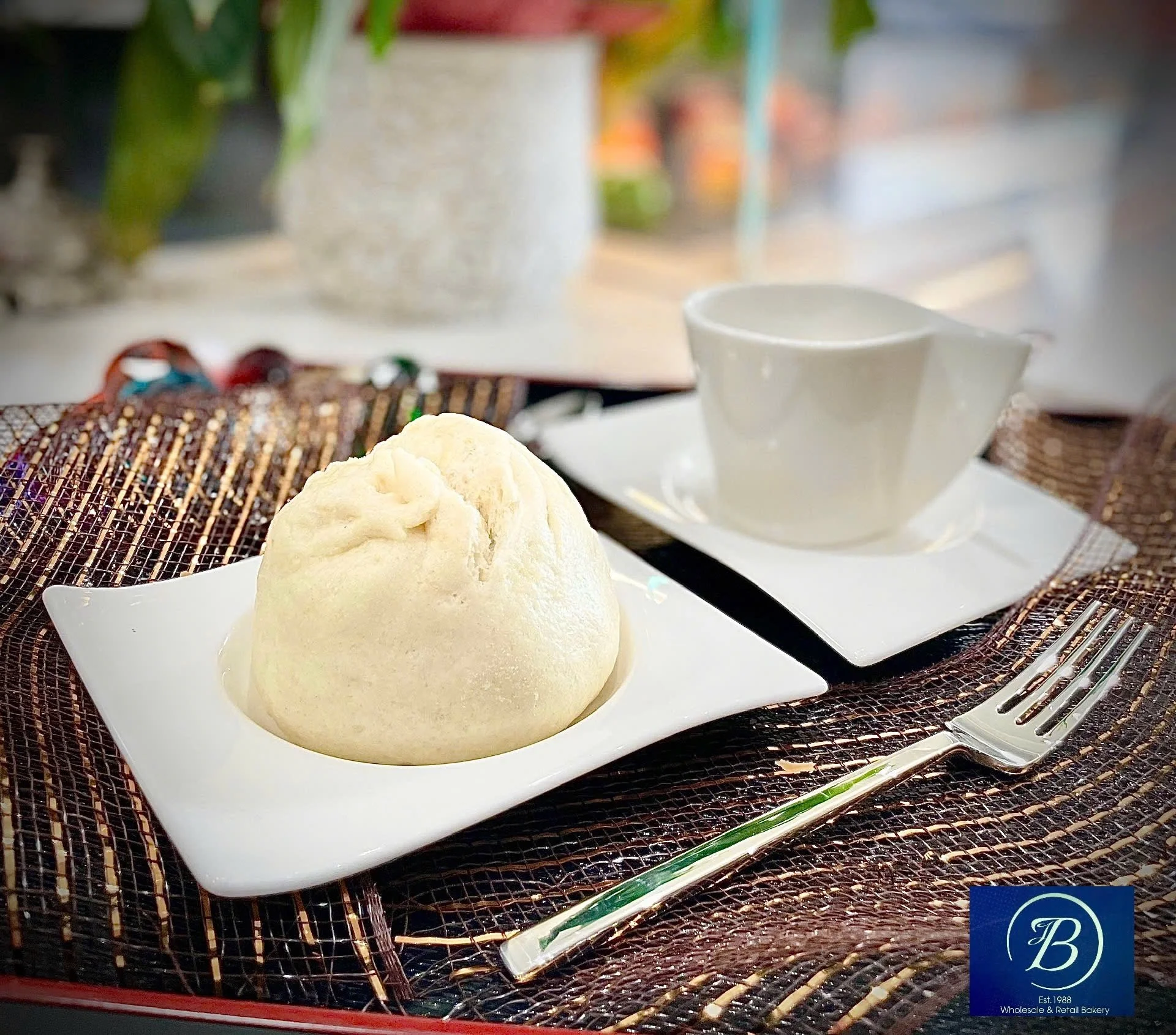 A white bowl with a mound of vanilla ice cream on a black wire rack, next to a white coffee cup with a saucer, silver spoon, and a clear plastic straw on a table with a decorative mat, in a bakery or cafe setting.