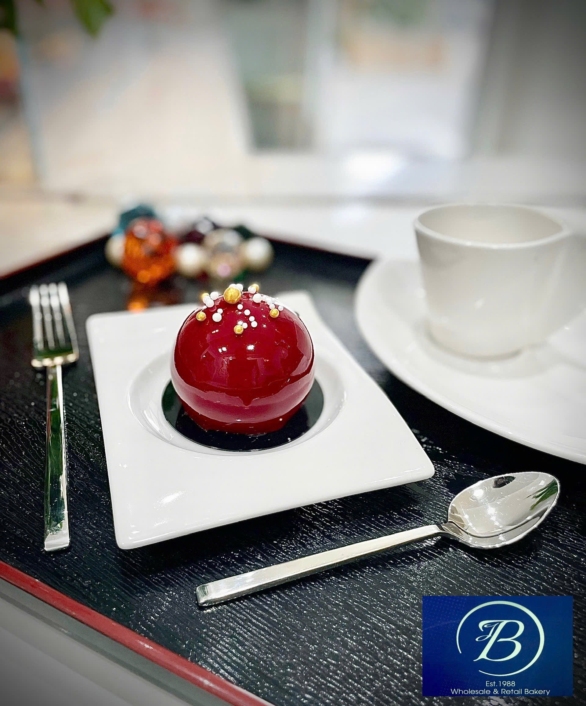 A red, dome-shaped pastry decorated with white, gold, and pink edible pearls on top, served on a white square plate. There are other assorted pastries visible in the background. The setting appears to be a bakery, with a blue logo indicating 'Est. 19