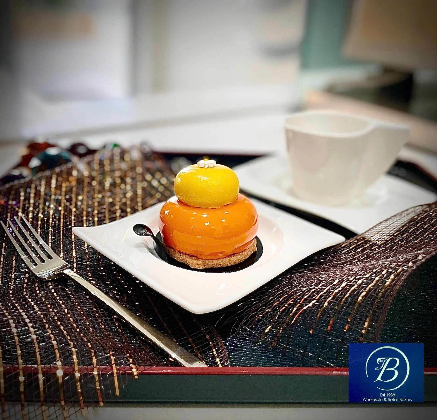 A layered pastry with yellow, orange, and red glossy fruit-like toppings, garnished with small white decorative pieces, served on a white plate with a silver spoon, on a table with woven placemats near a white coffee mug and a patterned brown cloth.