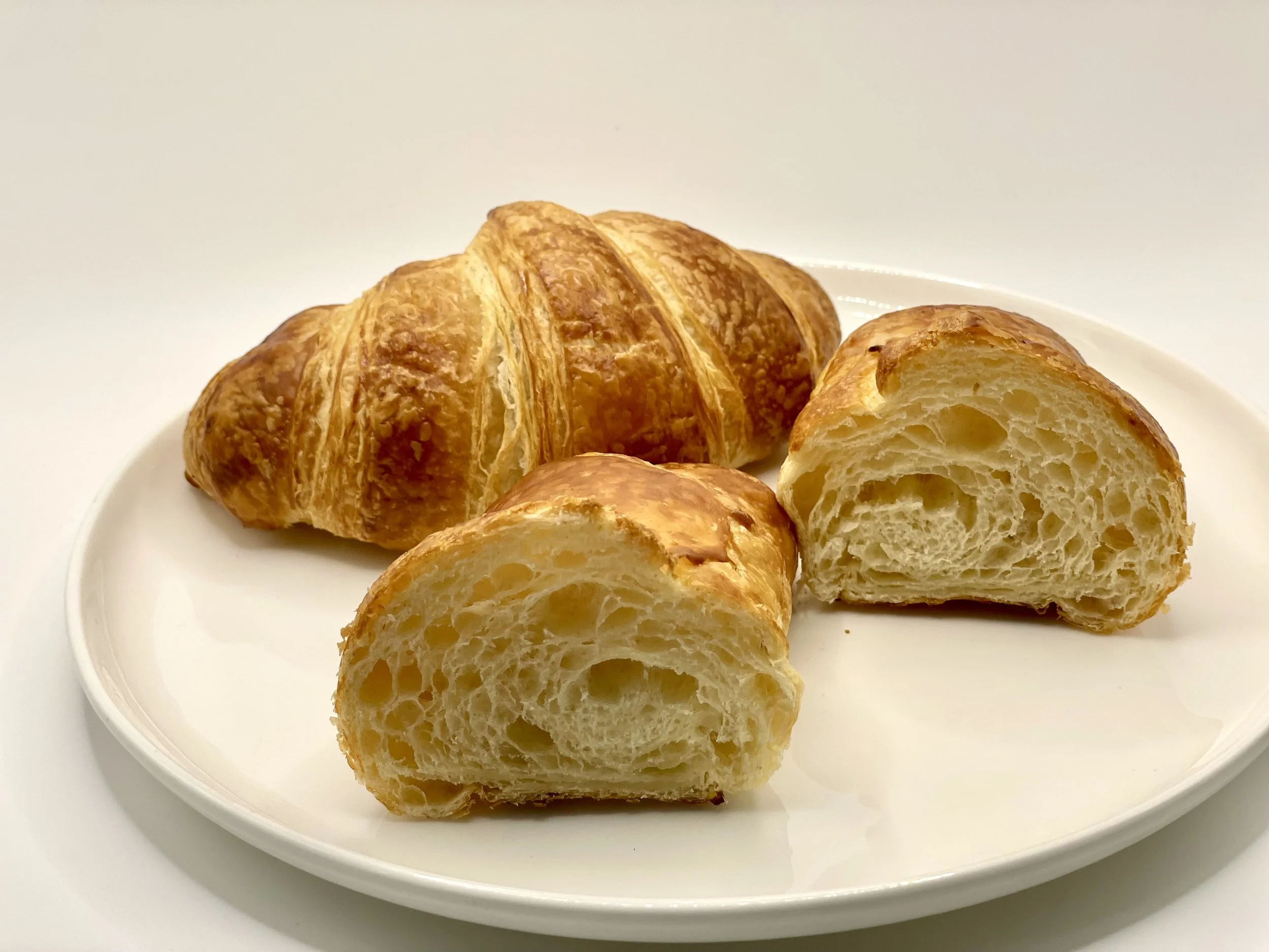 A white plate holding a croissant cut in half, showing its airy and flaky interior, with a plain white background.