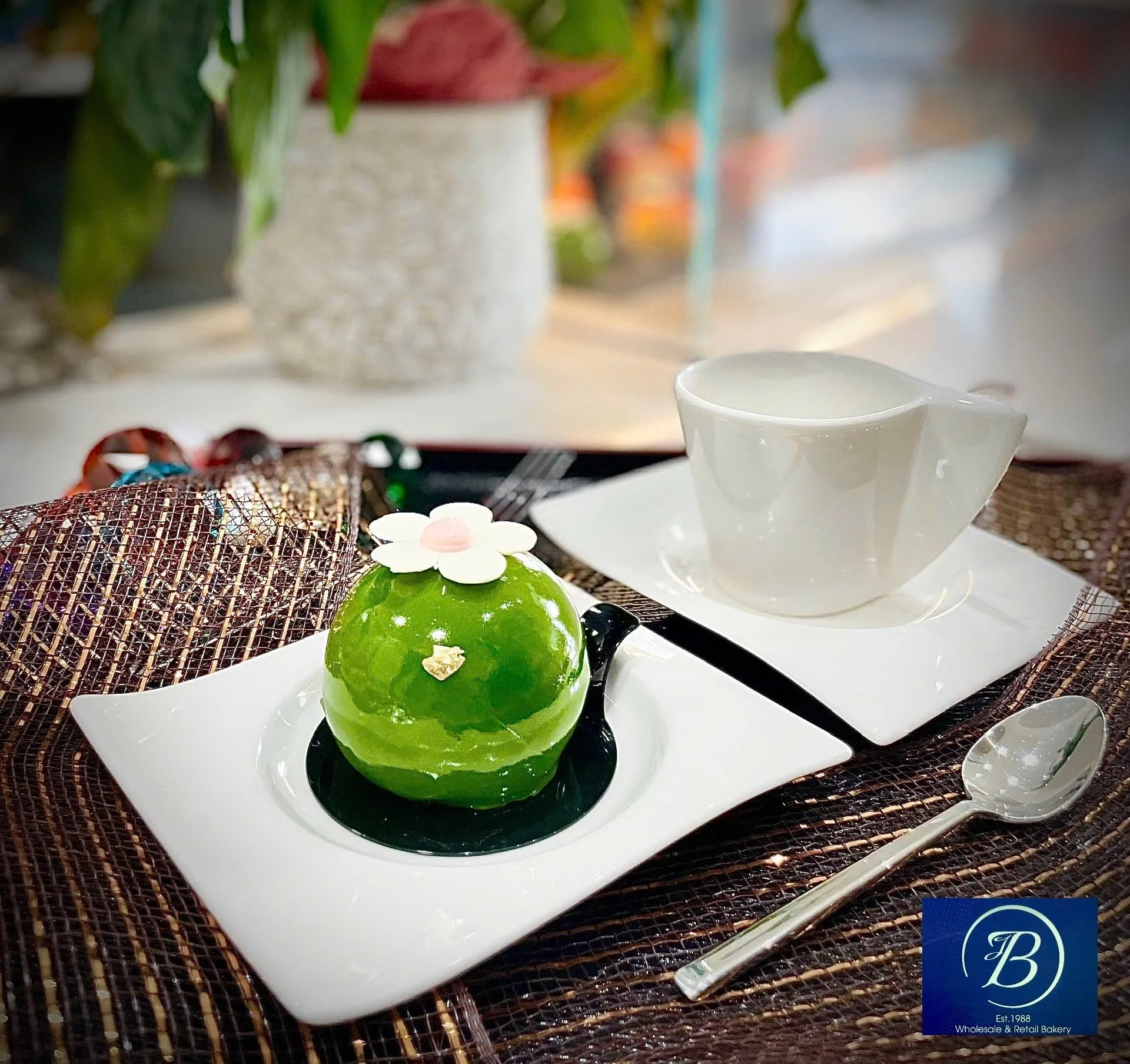 A white rectangular plate holding three green domed desserts topped with a white flower and a pink dot, placed on a woven placemat with some colorful wrapped candies and a white ceramic pitcher in the background.