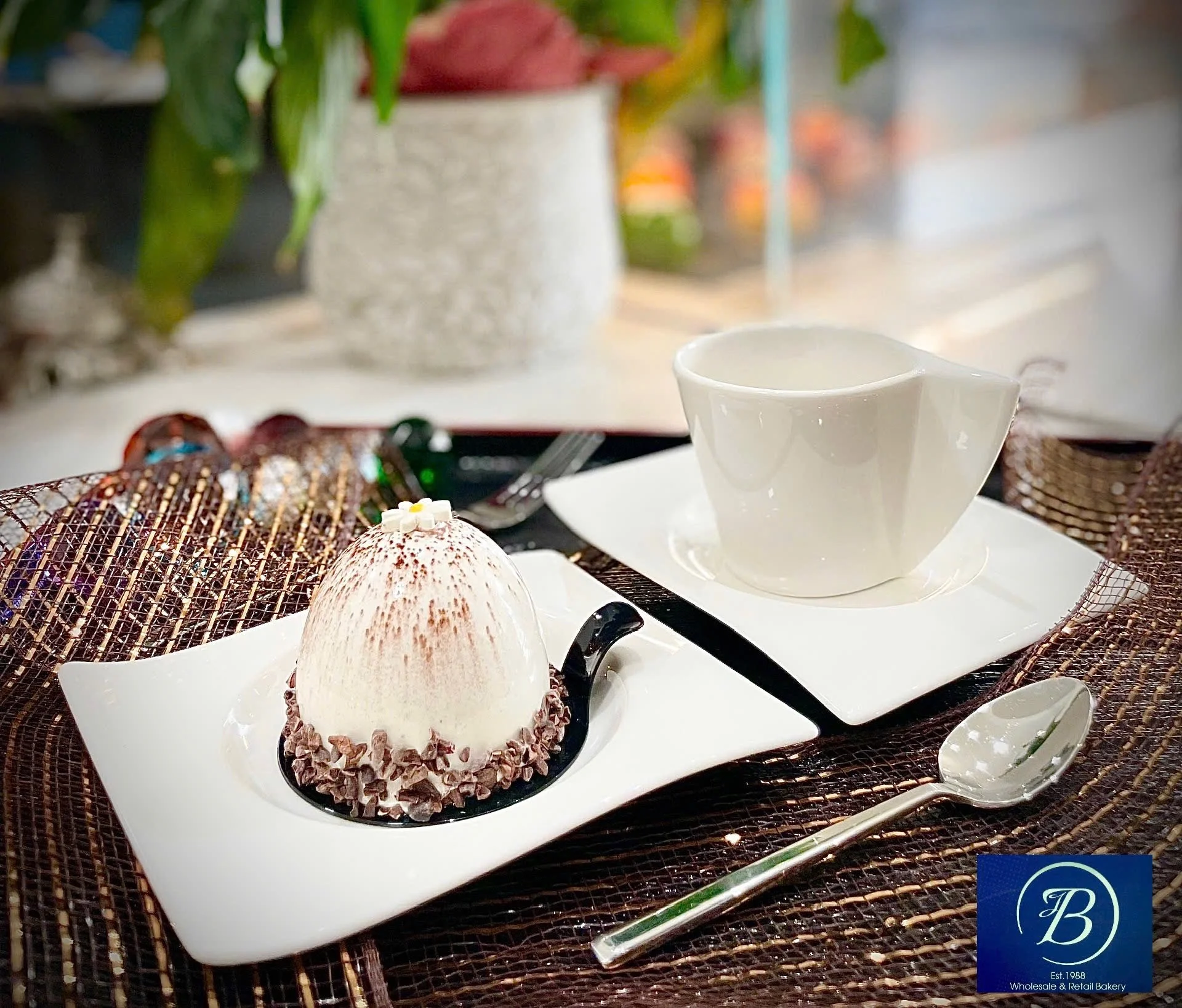 Assorted desserts on a white platter, including a round scoop of ice cream topped with chocolate shavings and a small white flower, surrounded by chocolate-dipped treats and a coffee cup in the background. The setting appears to be a bakery or café.