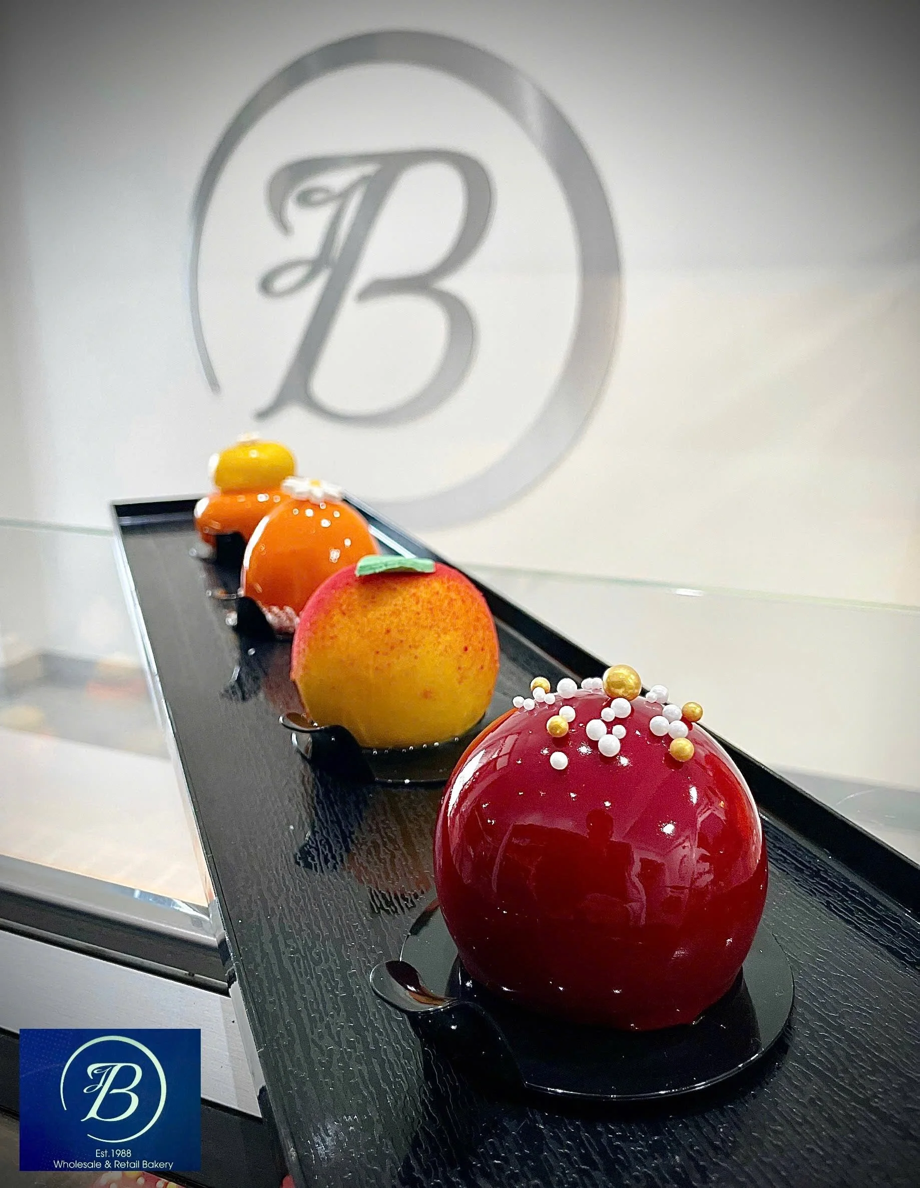 Five glossy, decorated, dome-shaped cakes on black trays, with botanical and pearl topping, placed on a metallic surface. Behind the cakes, a white wall features a gray logo with the letter 'B' encircled, and text indicating 'Est. 1988 Wholesale & Re