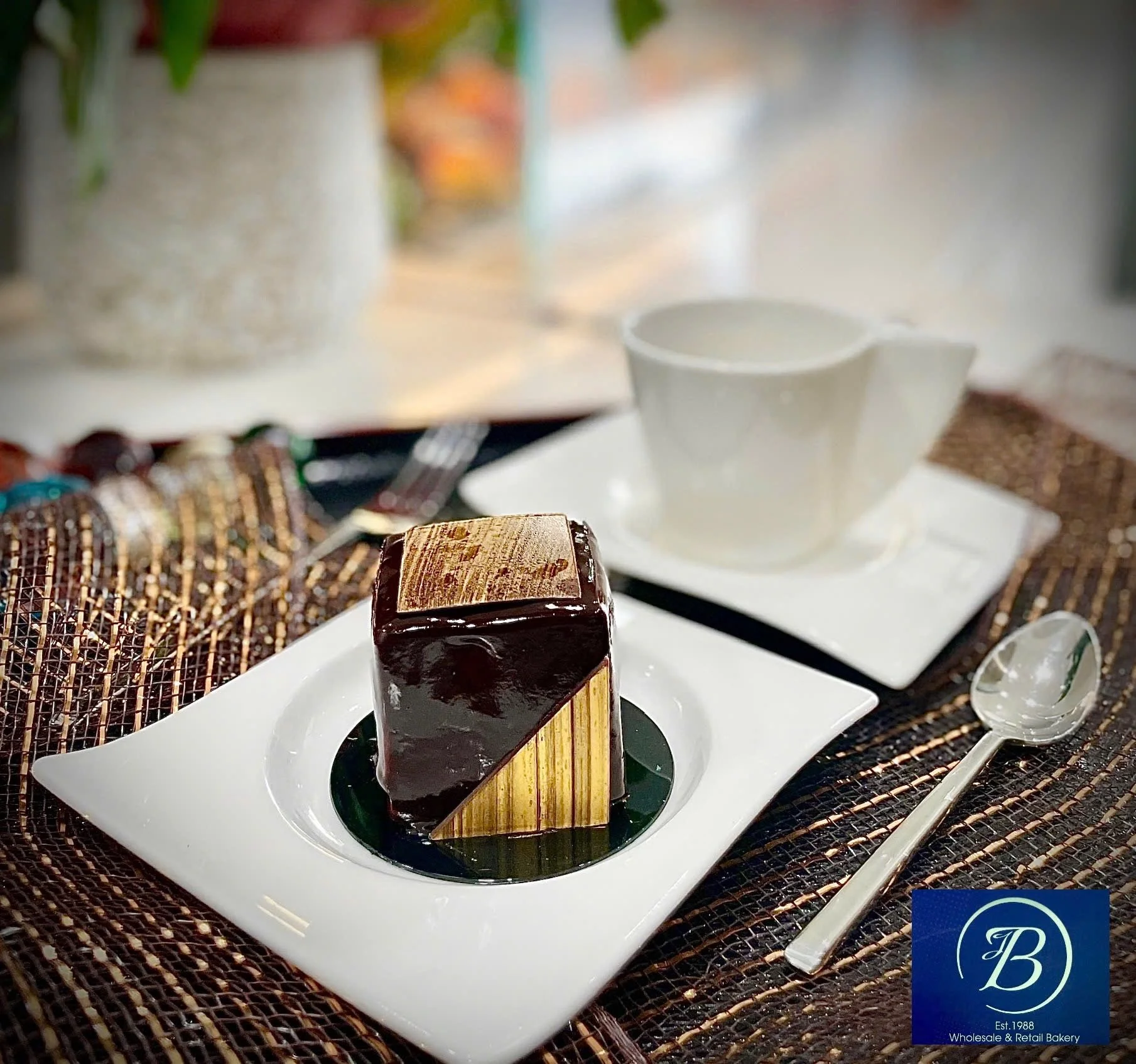 A dessert with a chocolate glaze and a decorative pattern on top, served on a white plate with a green garnish underneath. In the background, there is a white cup on a saucer, a spoon, and blurred decorative items on a table.