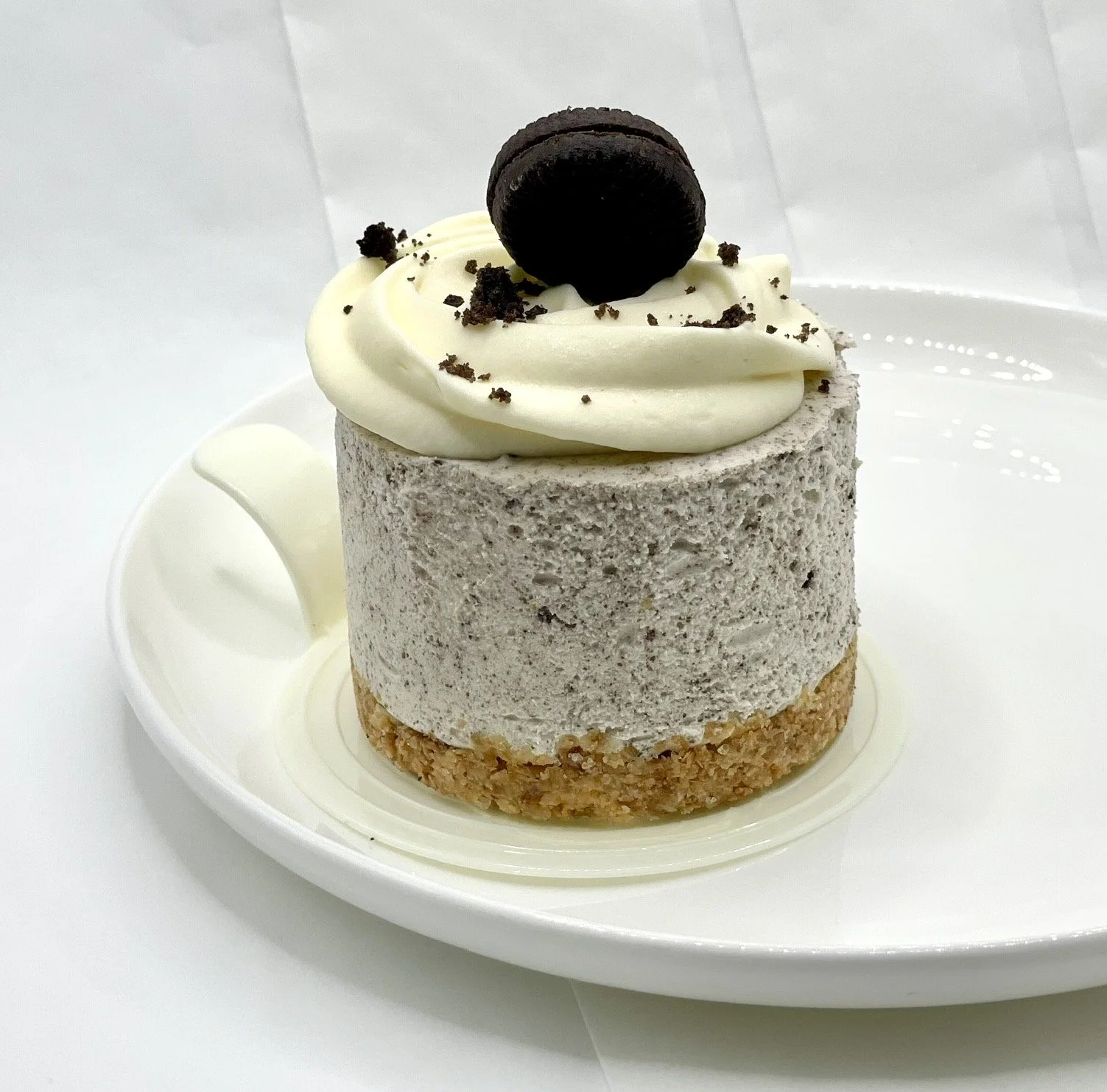 A dessert with a layered appearance on a white plate. The bottom layer is a crunchy biscuit crust, the middle layer is a light gray mousse, and the top is a swirl of white cream topped with a chocolate cookie and crumbles.
