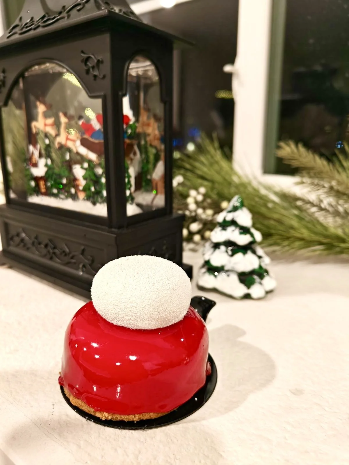 A festive Christmas display with a red glossy Christmas cake topped with a white snowball decoration, a black lantern with miniature Christmas figurines inside, a small snow-covered Christmas tree, and pine branches with white berries near a window.