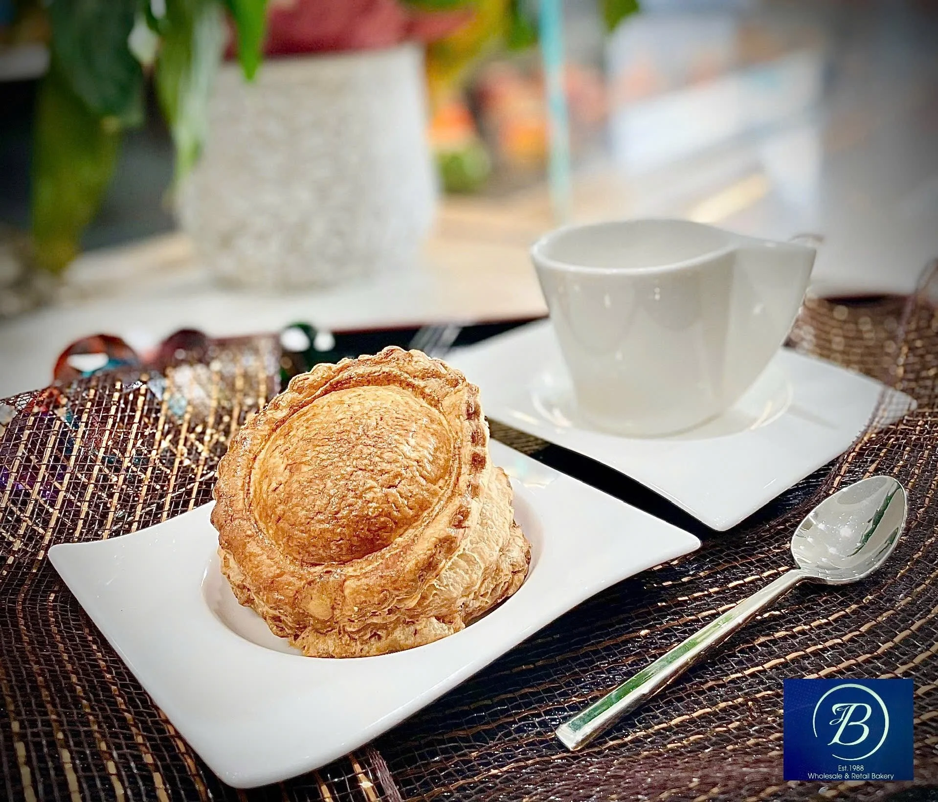 Close-up of a flaky, golden croissant on a white rectangular plate, placed on a dark woven placemat. In the background, a white cup, some plants, and a blurred view of a home or café interior are visible.