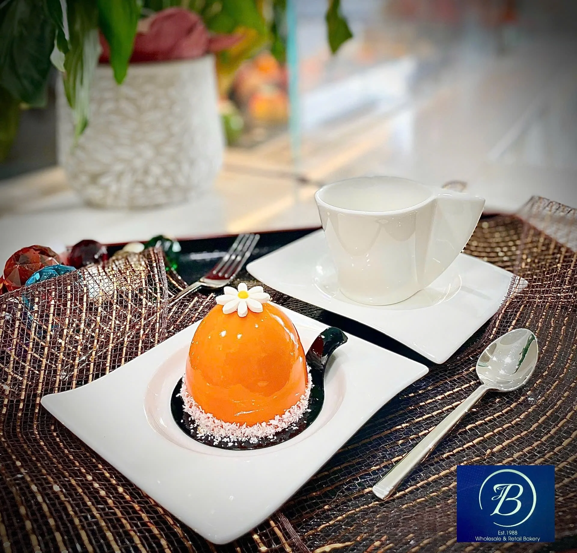 A white square plate holds an orange mousse dome cake decorated with a white flower and small white spots, surrounded by shredded coconut on the plate's edges. In the background, other desserts are partially visible, and silver utensils are placed on