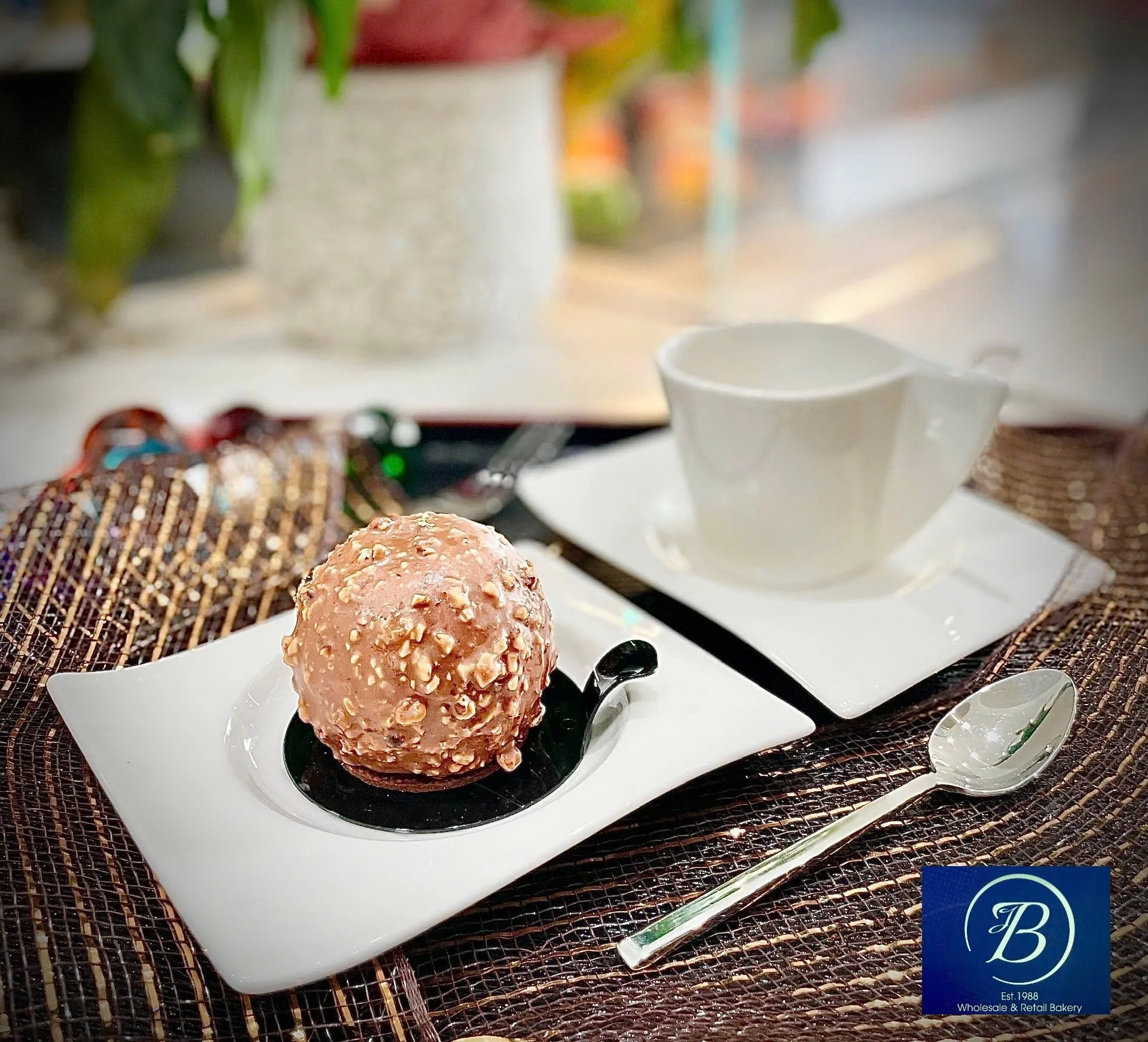 A scoop of pink ice cream with nuts on top served in a white dish, placed on a woven placemat with a spoon next to it, and a white teacup and saucer in the background, on a table set for dessert.