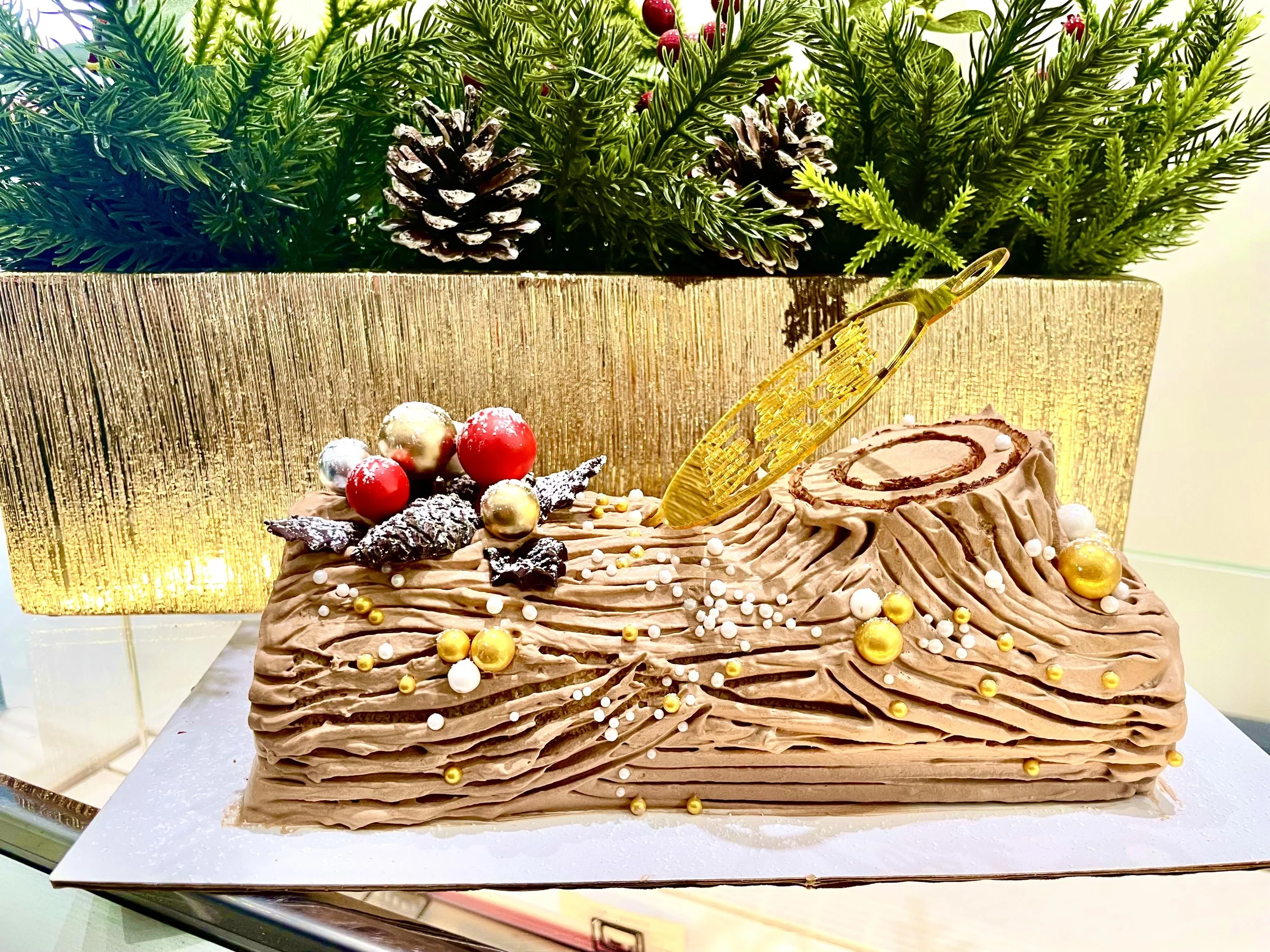 A chocolate Christmas cake decorated with white and gold edible pearls, holiday-themed ornaments, and a gold decorative piece on top, with a gold and green garland and pinecones in the background.