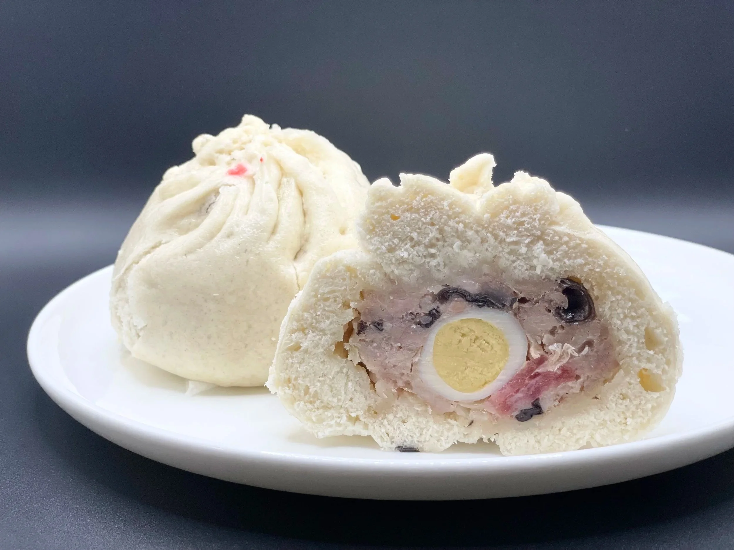 Close-up of a Chinese steamed bun with a soft texture, filled with black beans, ham, and a yellow egg center, served on a white plate against a dark background.